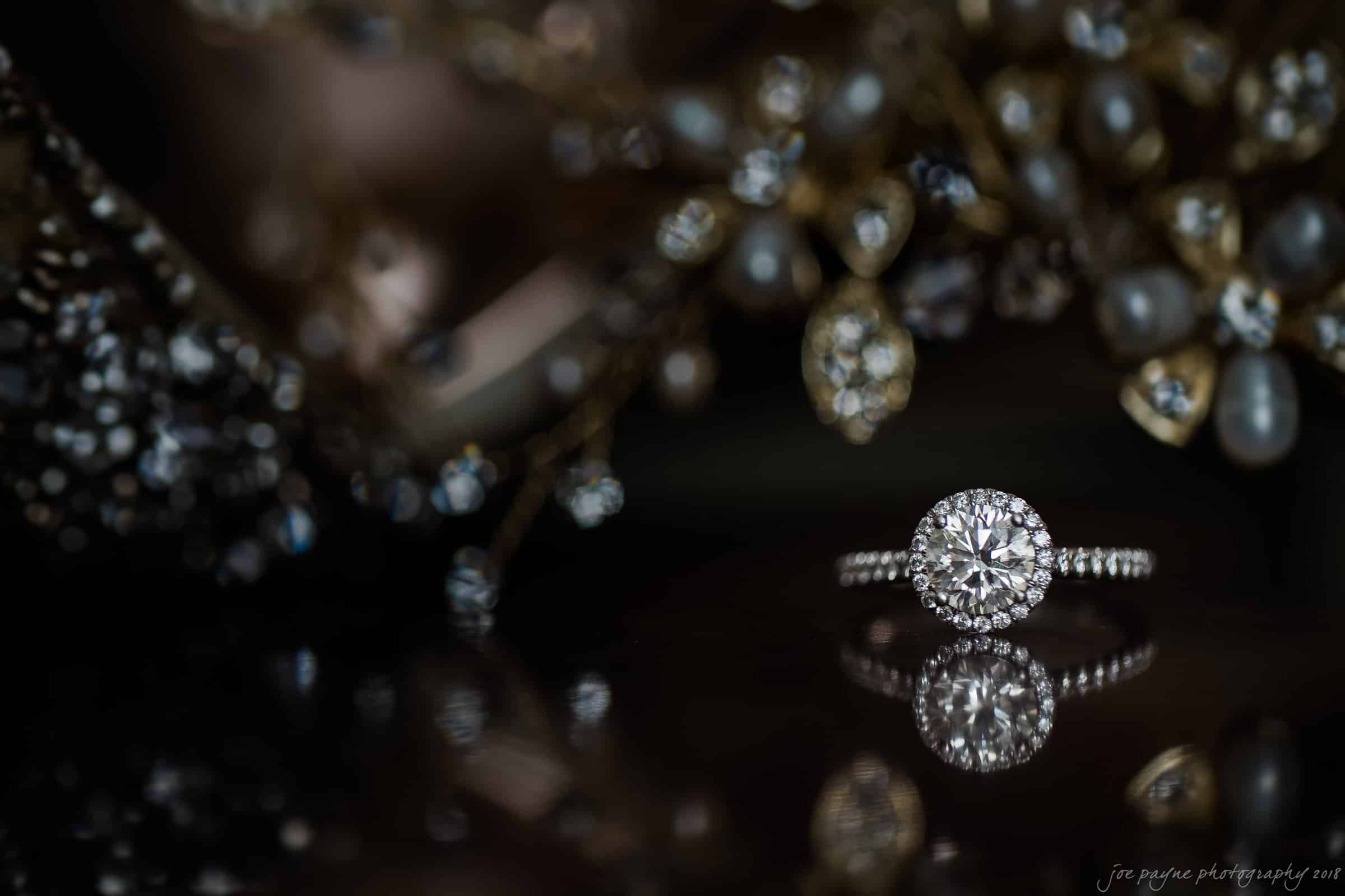 A diamond ring with a halo setting sits elegantly on a reflective surface. The background features blurred, sparkling elements that enhance the rings brilliance.
