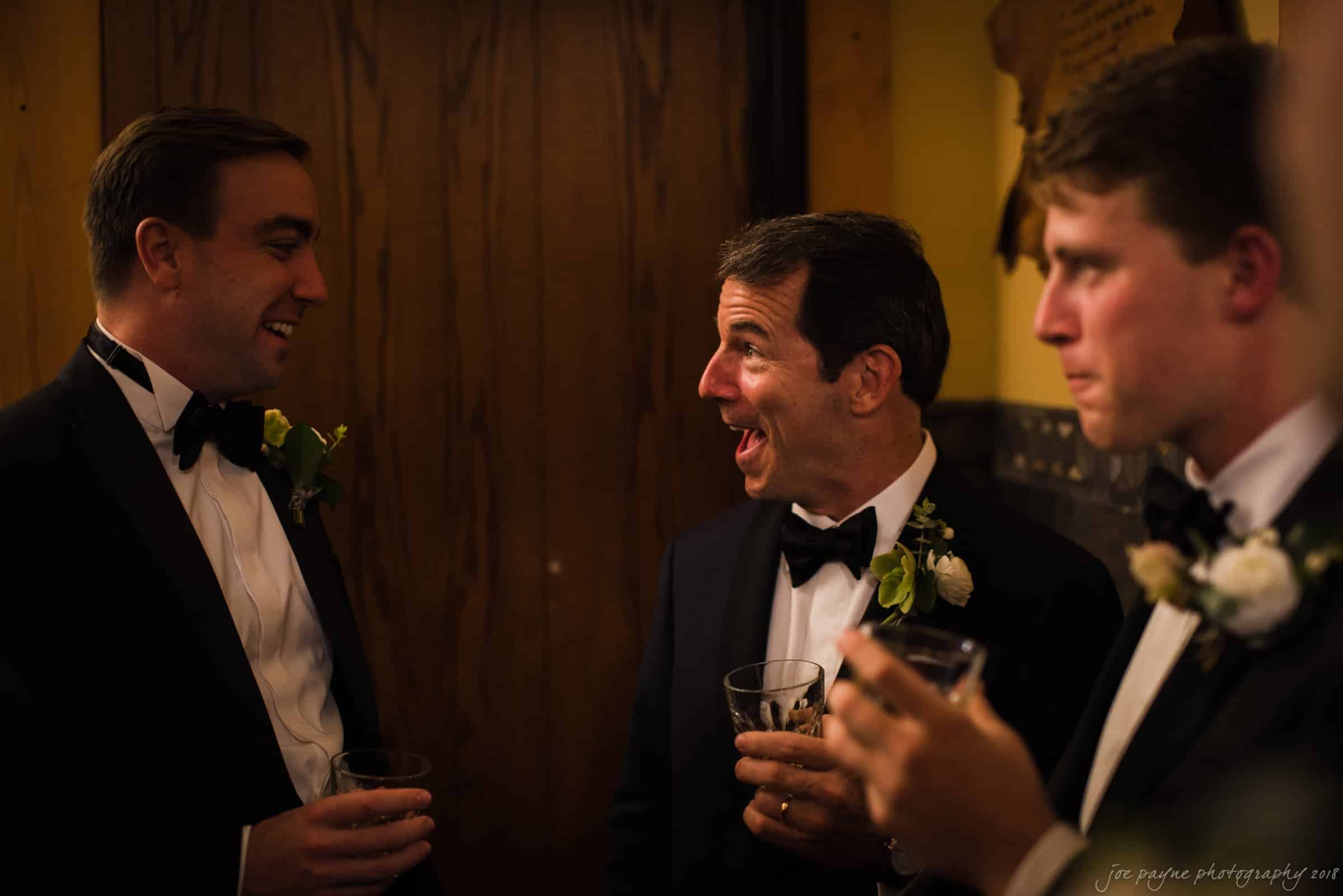 Three men in suits and bow ties, each holding a drink, are engaged in conversation. The man in the middle appears animated and smiling. They are in an indoor setting with warm lighting.