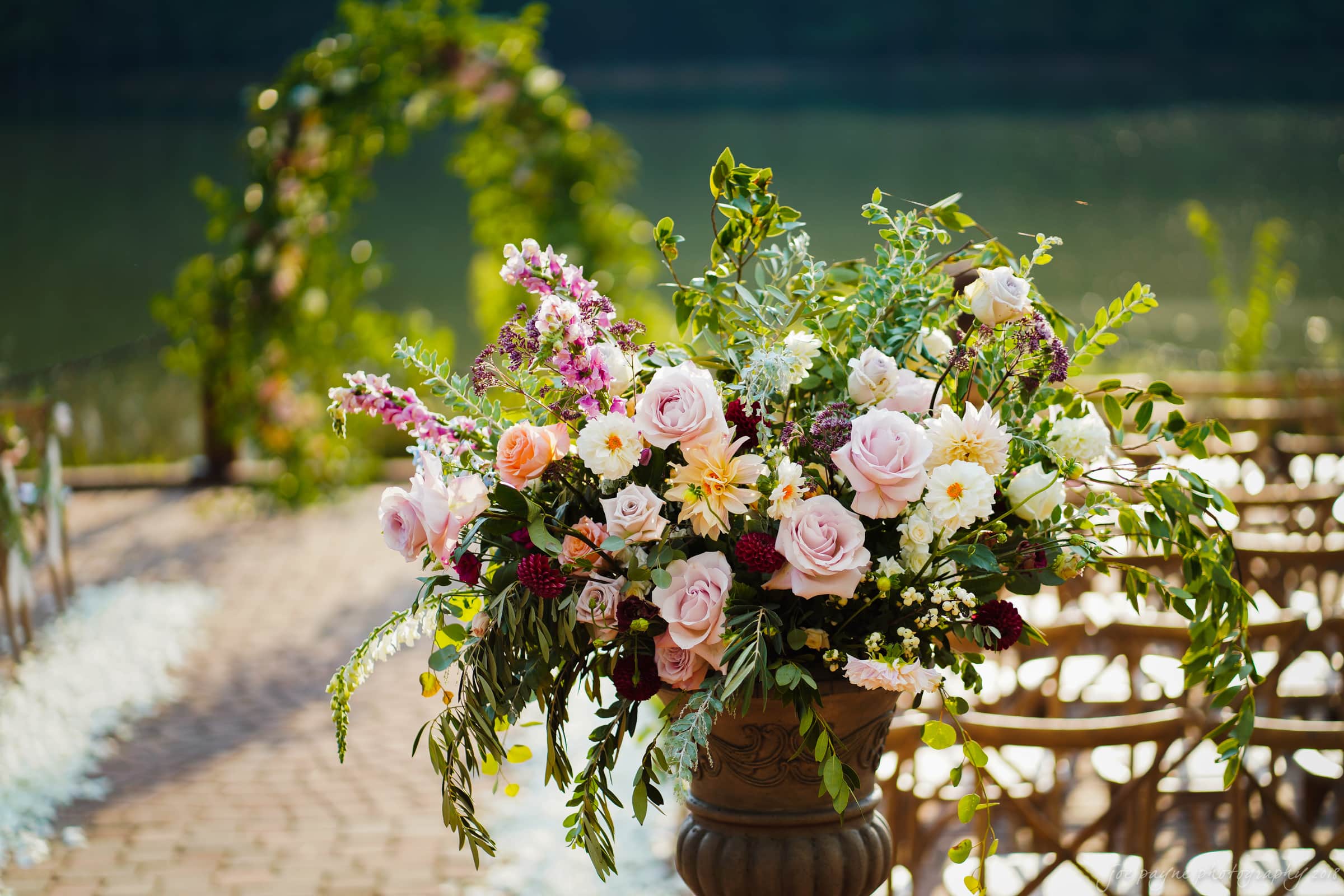 A lush floral arrangement with roses, daisies, and greenery in a vase stands near a pathway lined with chairs. An archway adorned with flowers is in the background, all set against the backdrop of a lake, creating a romantic outdoor setting.