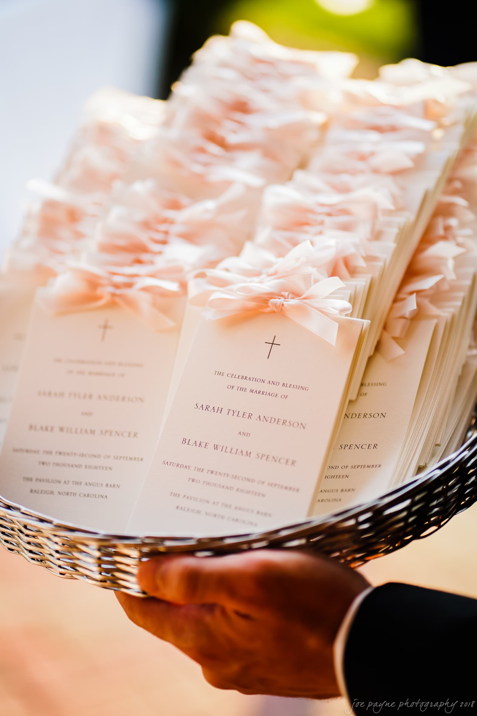 A basket filled with wedding programs decorated with light pink bows. The programs are held by a person in black attire. They include details about the marriage of Sarah Tyler Anderson and Blake William Spencer.