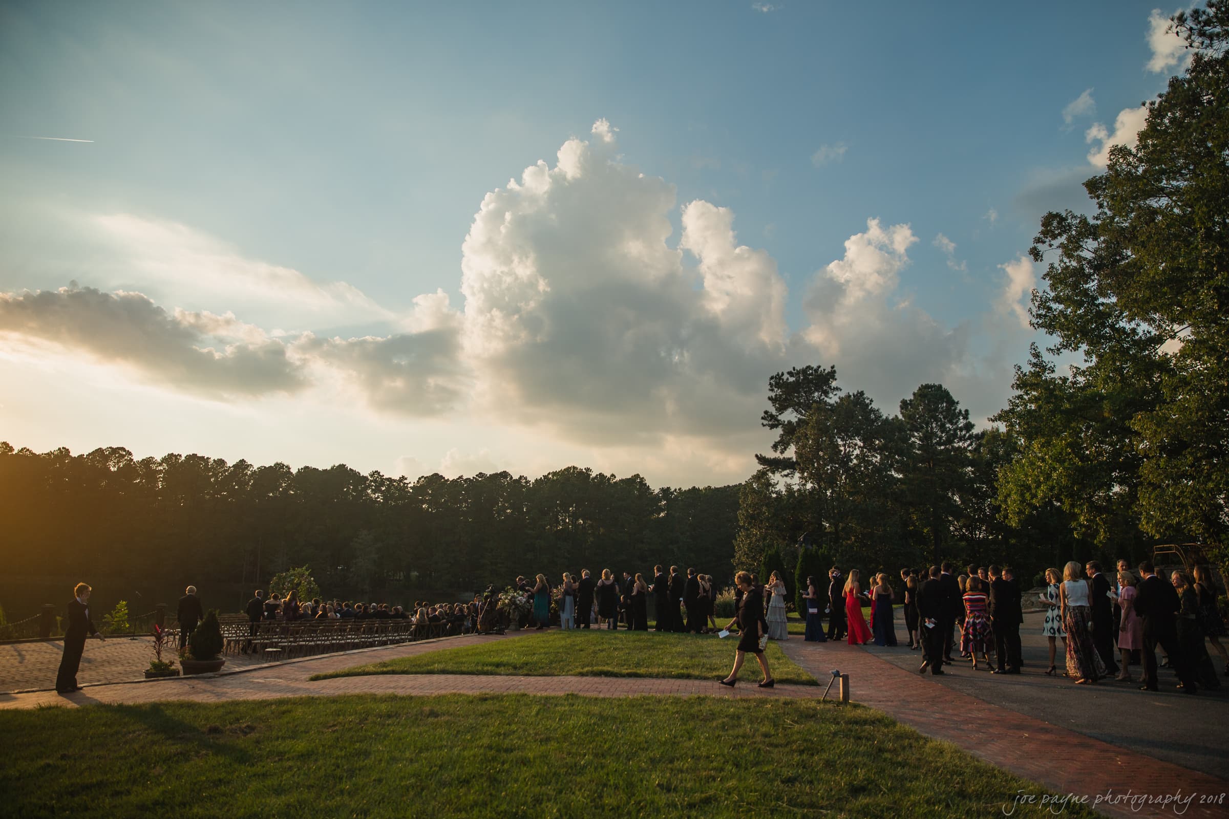 A large outdoor gathering with people wearing formal attire. The event is set against a backdrop of a forest with a sunset sky. The clouds are scattered and the sunlight casts long shadows on the ground.