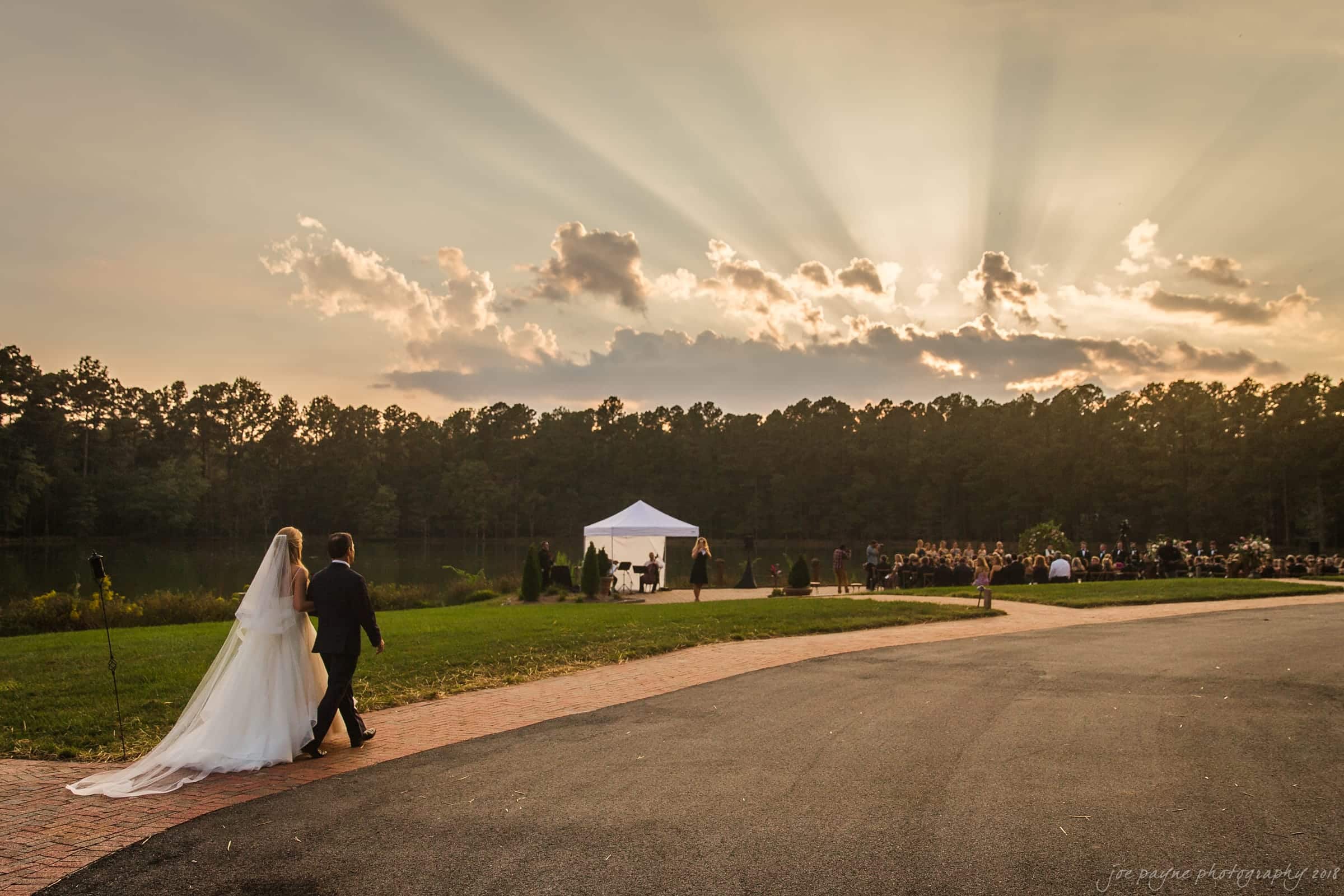 A bride and groom walk hand in hand toward an outdoor wedding ceremony by a lake. The sky is dramatic with sun rays streaming through clouds, and a canopy tent is set up with guests seated nearby. Trees and water create a serene backdrop.