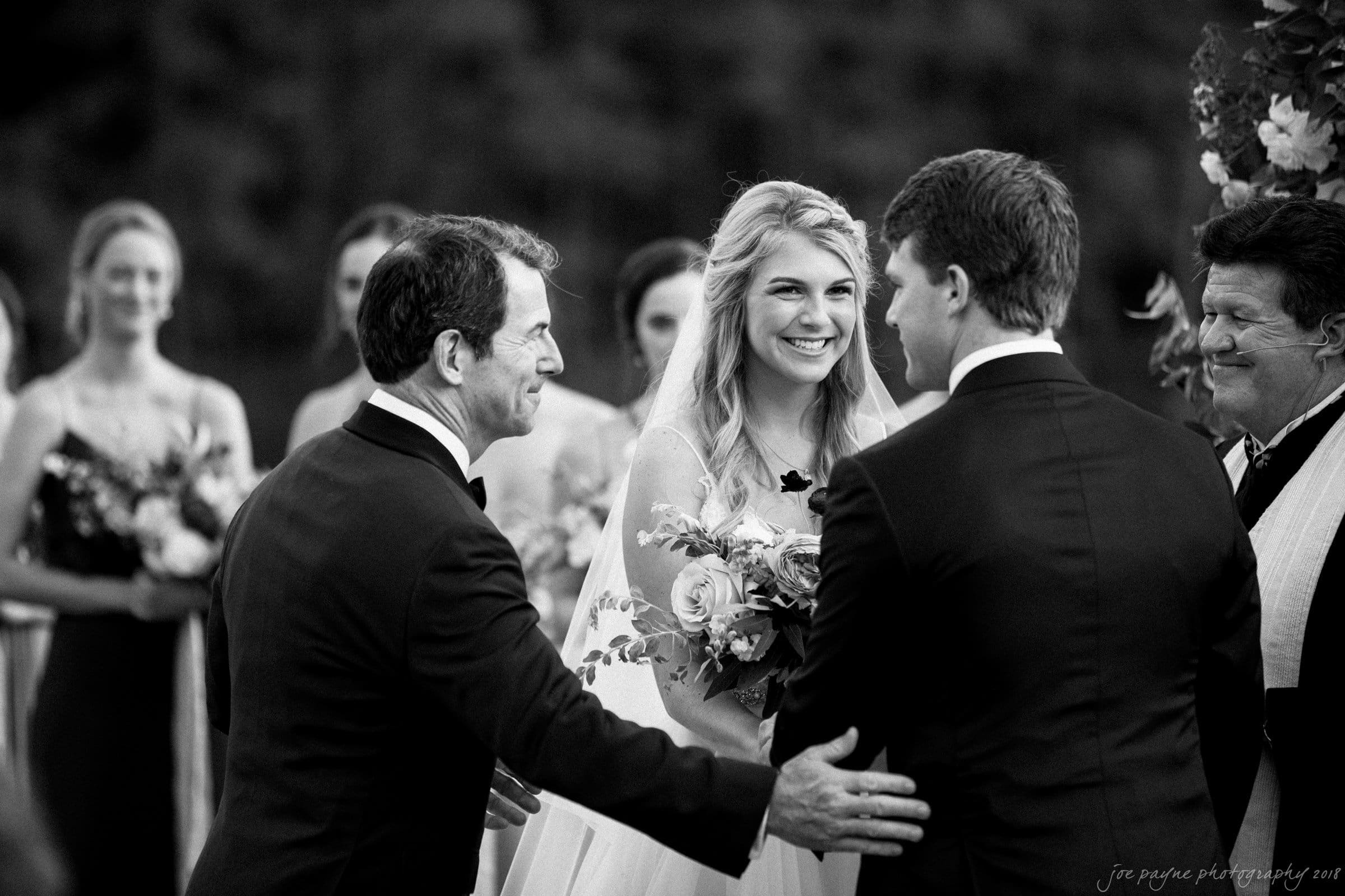 A bride smiling as her father gives her away to the groom during an outdoor wedding ceremony. Bridesmaids stand in the background holding bouquets. The scene is captured in black and white.