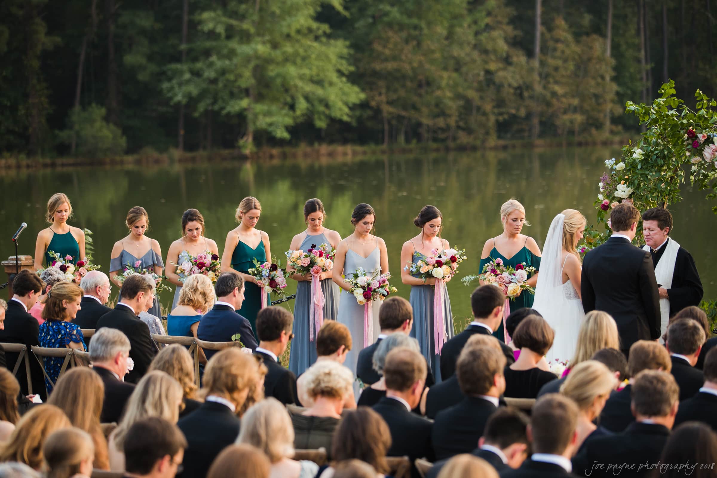 A wedding ceremony by a lakeside. The bride and groom stand before a minister, with bridesmaids in matching dresses holding bouquets beside them. Guests are seated, witnessing the event surrounded by lush greenery and calm water.