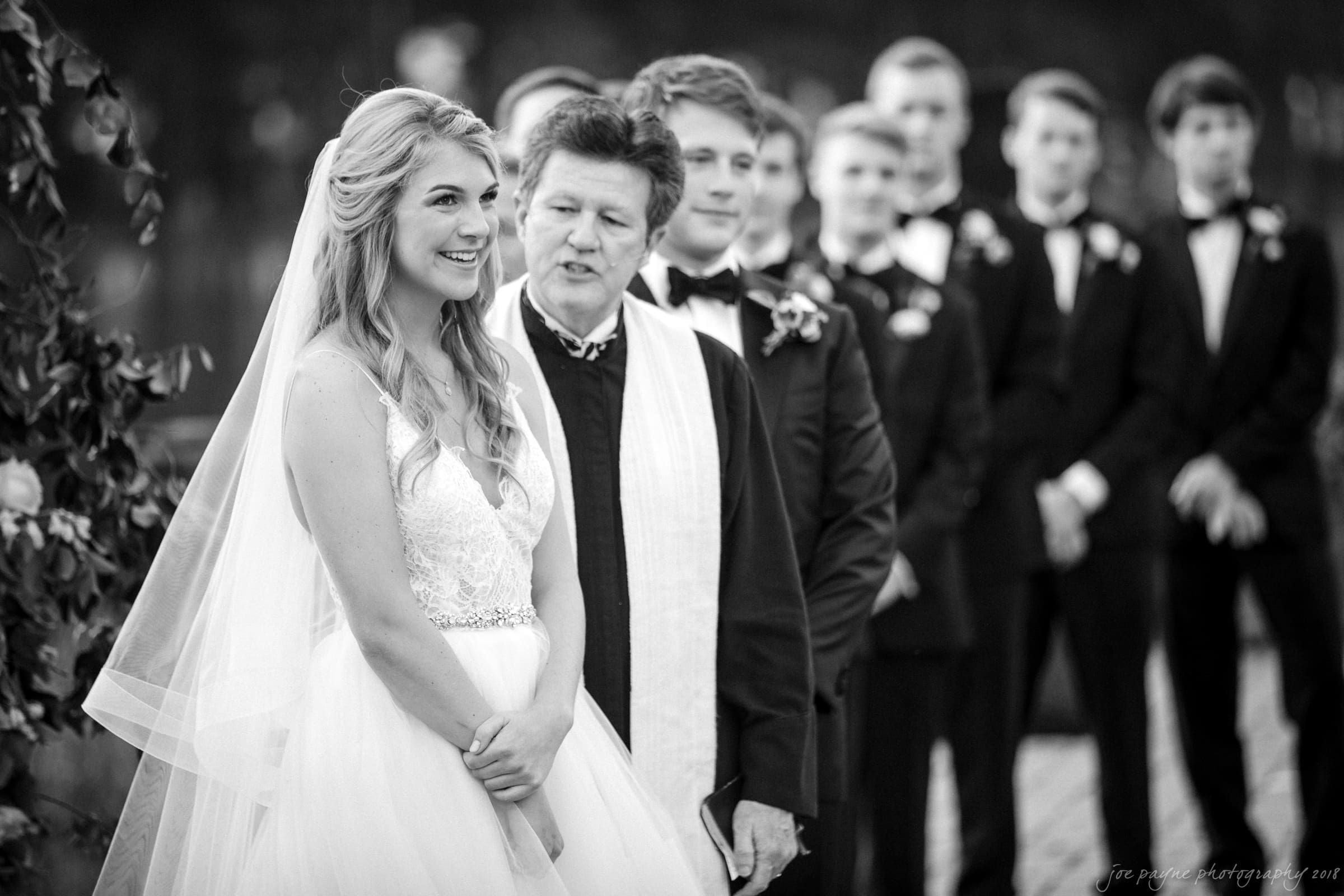 A black-and-white photo of a bride and officiant during a wedding ceremony. The bride is smiling, wearing a veil and a lace dress. Groomsmen in suits stand in the background. The scene is outdoors with a soft focus.