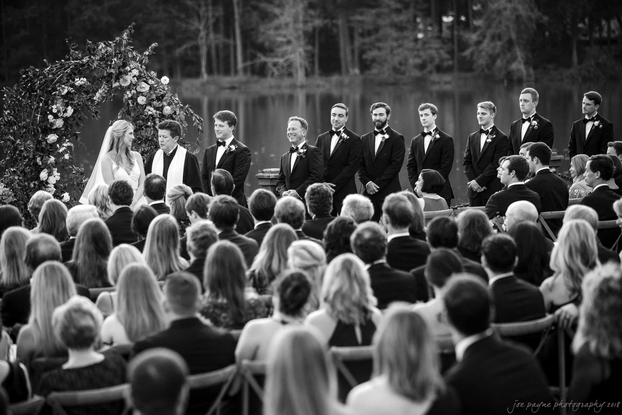 Black and white photo of a wedding ceremony by a lake. The bride and groom are standing under a floral arch with a minister. Groomsmen in tuxedos stand nearby. Guests are seated, facing the ceremony. Trees are visible in the background.