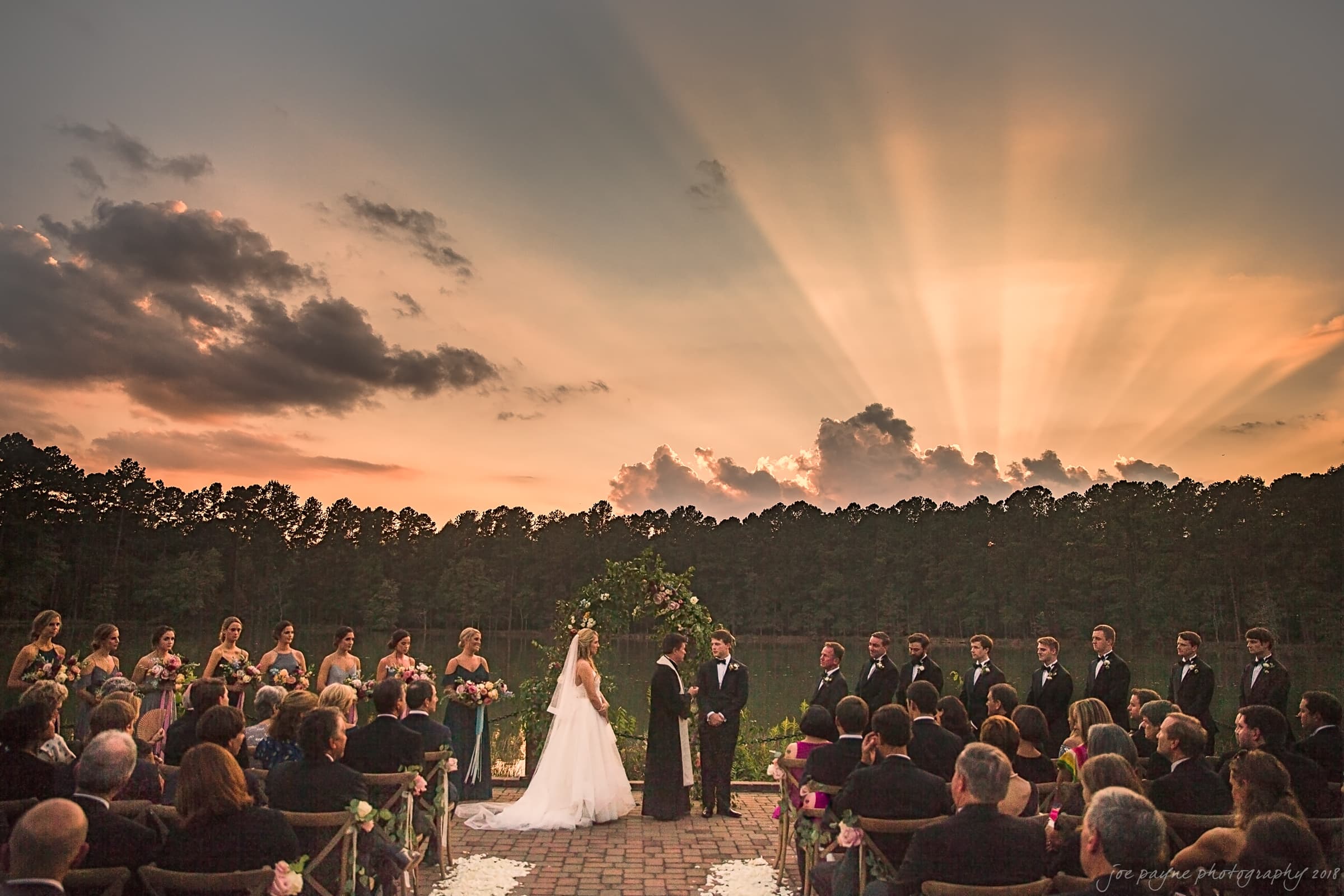 A bride and groom stand at an outdoor altar during sunset. Bridesmaids and groomsmen are lined up beside them, with guests seated in the foreground. The sky is colorful with dramatic rays of light and clouds.