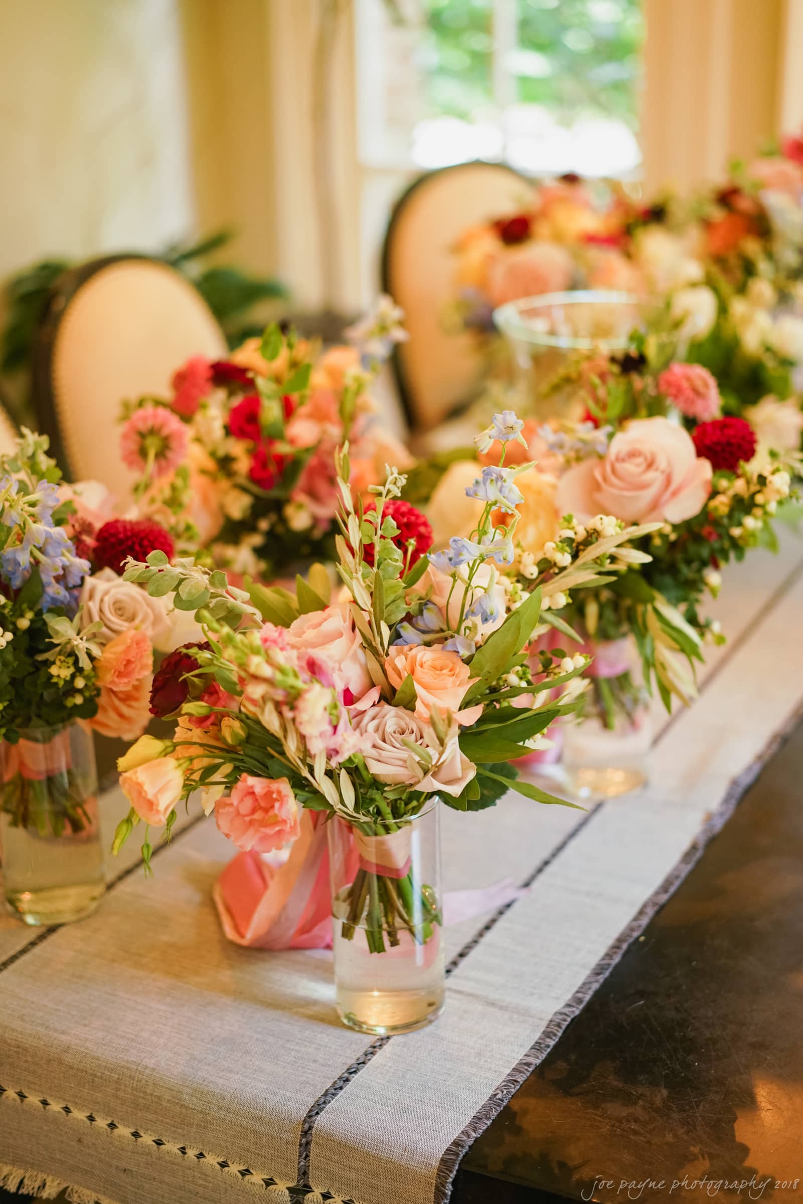 A table is elegantly decorated with bouquets of colorful flowers, including roses and greenery, arranged in vases. The table is covered with a light tablecloth, and the setting has a soft, welcoming atmosphere.