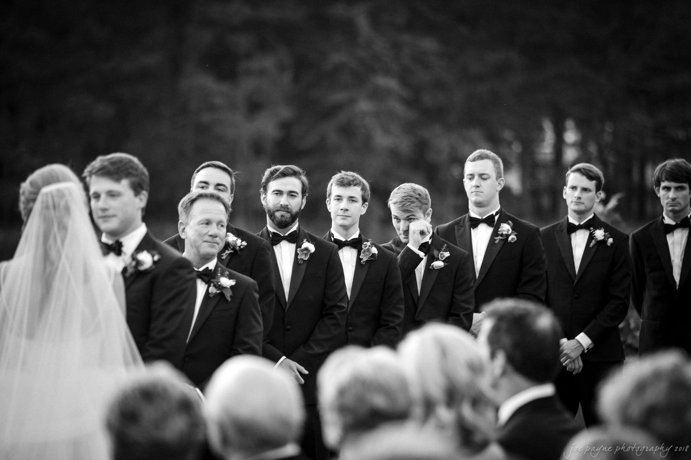 A black-and-white photo of a wedding ceremony shows a bride with a veil facing a line of groomsmen in tuxedos. They stand outdoors, with trees in the background, as they watch the ceremony intently.