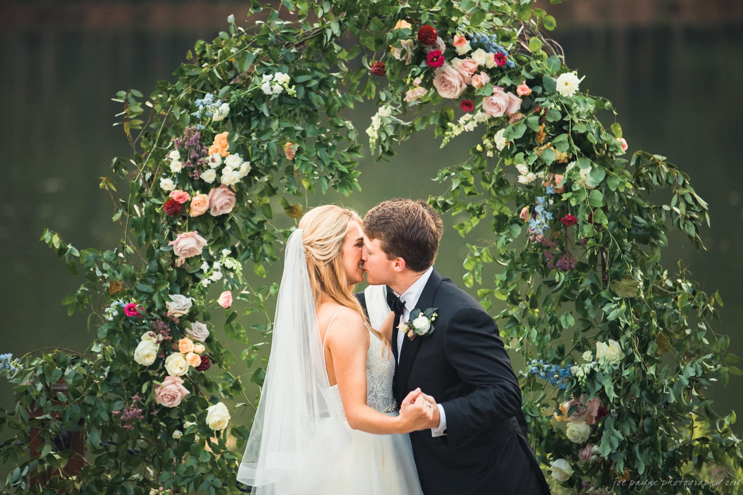 A couple in wedding attire shares a kiss beneath a floral arch adorned with greenery and colorful flowers, set against a serene outdoor backdrop. The bride wears a veil and a white dress, and the groom is in a black suit with a boutonniere.