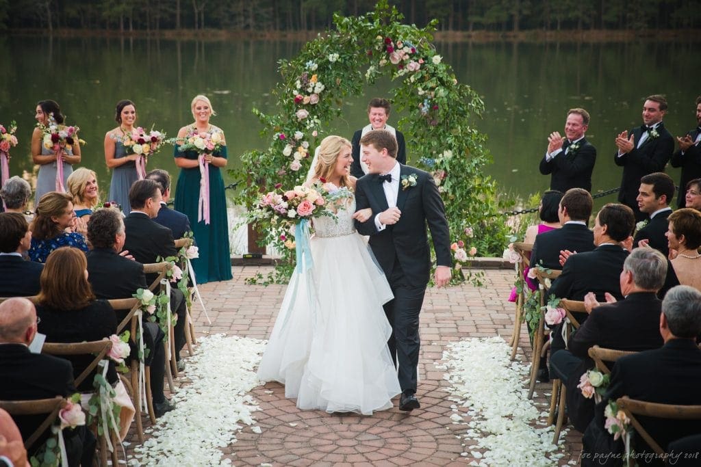 A bride and groom joyfully walk down the aisle after their outdoor wedding ceremony by a lakeside. Bridesmaids and groomsmen hold flowers in the background. Guests are seated and clapping on both sides amidst scattered white petals.