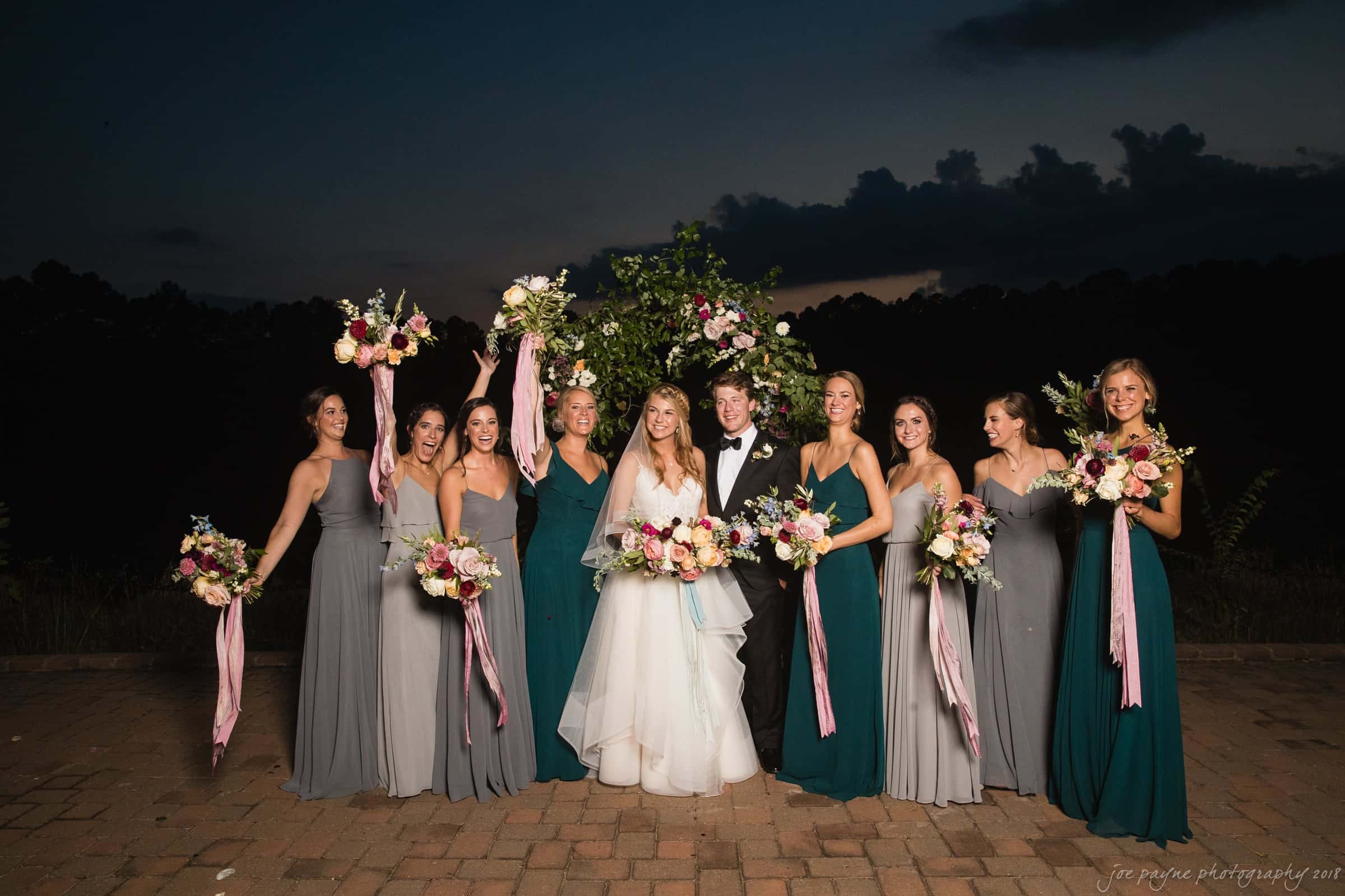 A wedding party poses outdoors during the evening. The bride and groom are surrounded by bridesmaids in blue and gray dresses, all holding bouquets. A floral arch is in the background against a dimly lit sky.