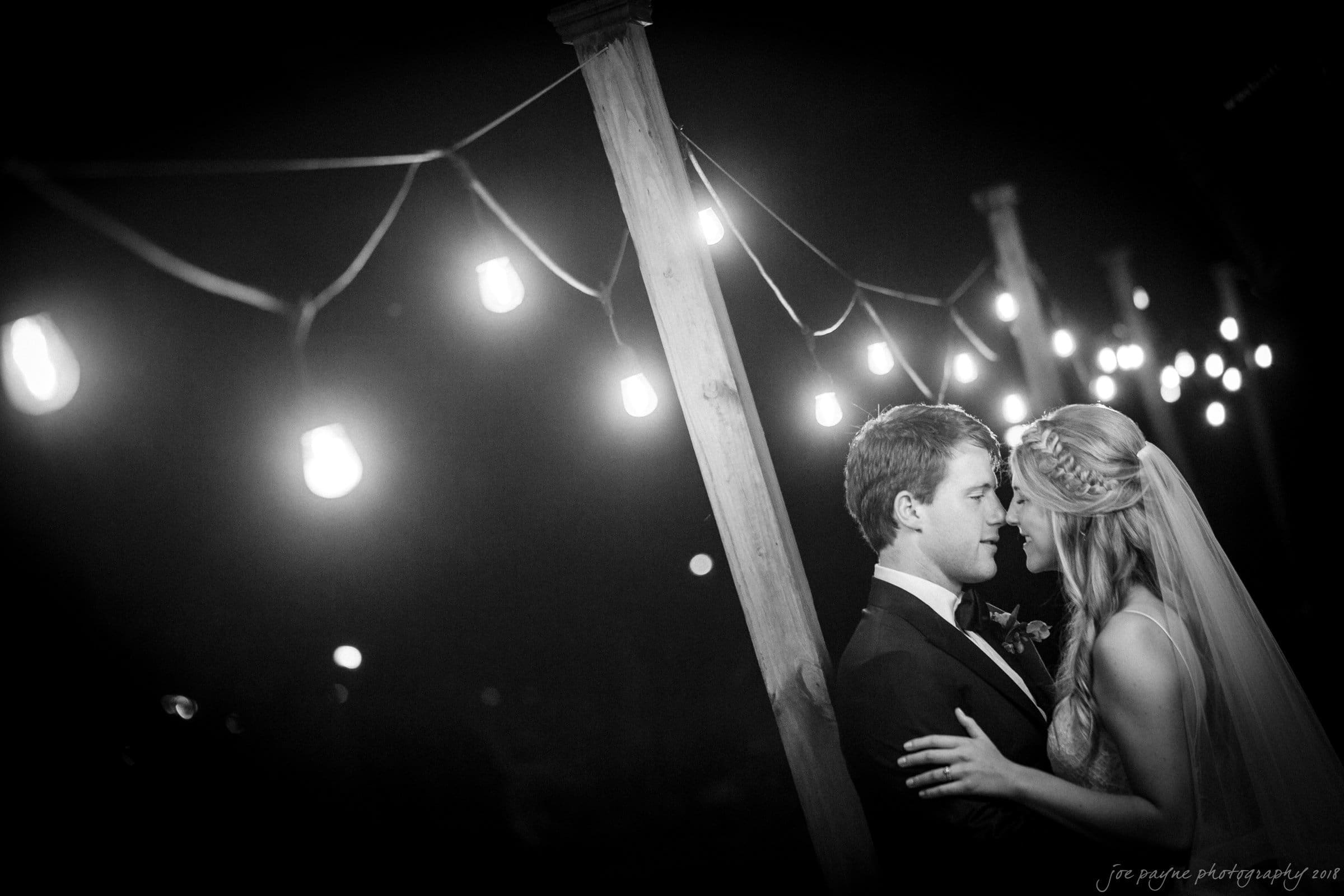 A black and white photo of a couple embracing in a romantic setting, surrounded by string lights. The bride wears a veil and headpiece, while the groom is in a suit. The atmosphere is intimate and serene.