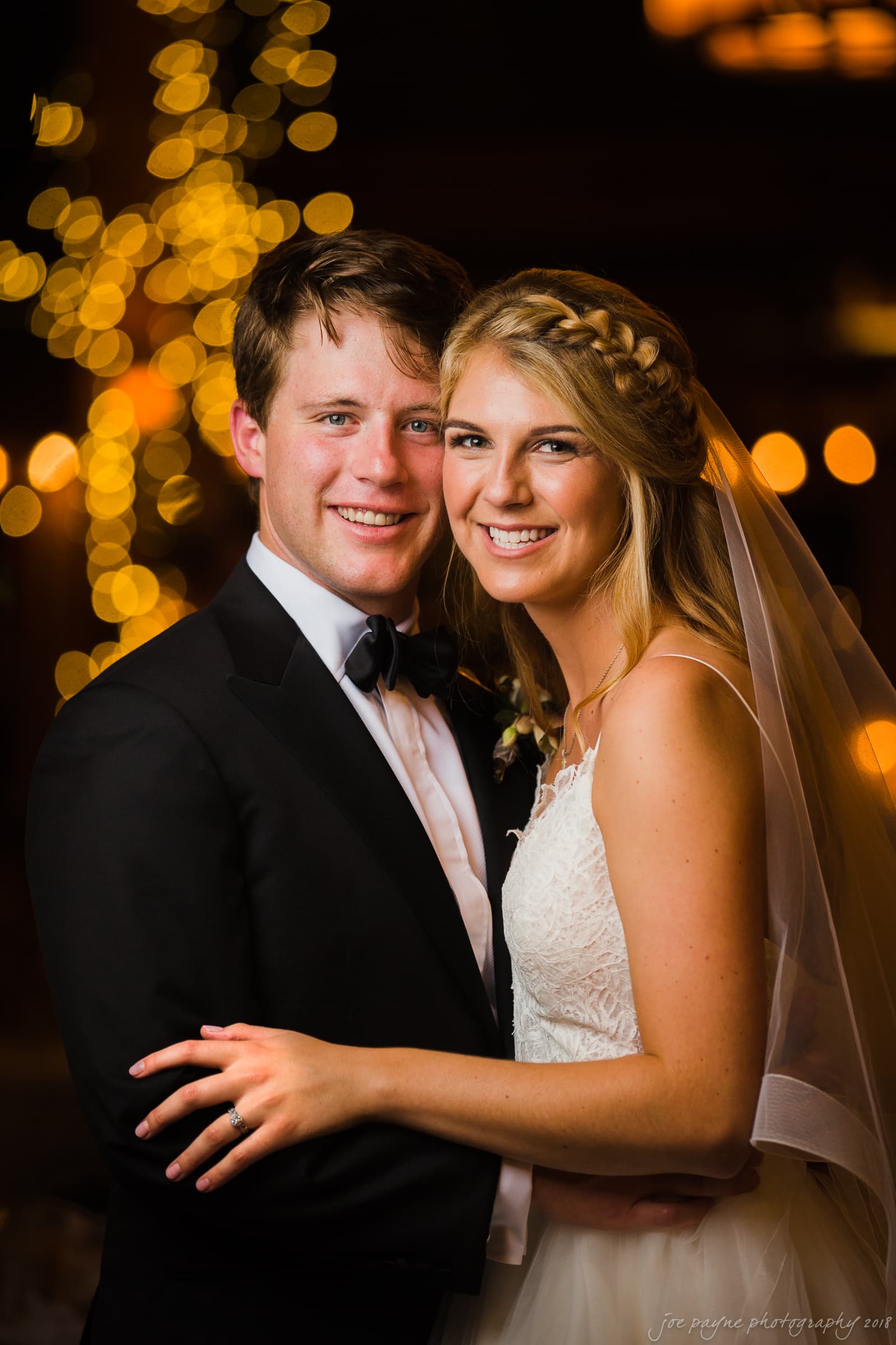 A bride and groom smile and embrace at their wedding, dressed in formal attire. The bride wears a white gown and veil, while the groom wears a black suit. Warm, blurred lights twinkle in the background, creating a romantic atmosphere.