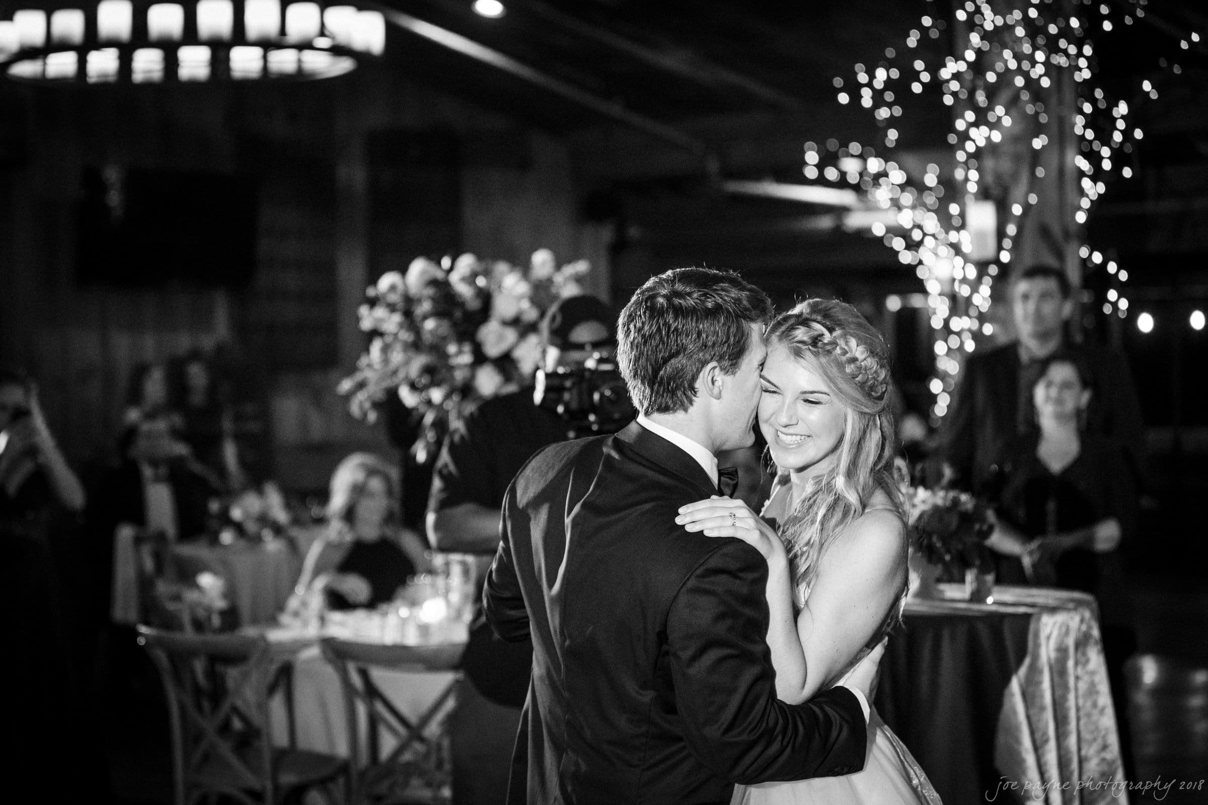 A black and white photo of a couple dancing in a warmly lit venue. The woman smiles while holding the mans shoulders, and he faces her. In the background, guests sit at tables decorated with flowers, and string lights twinkle.