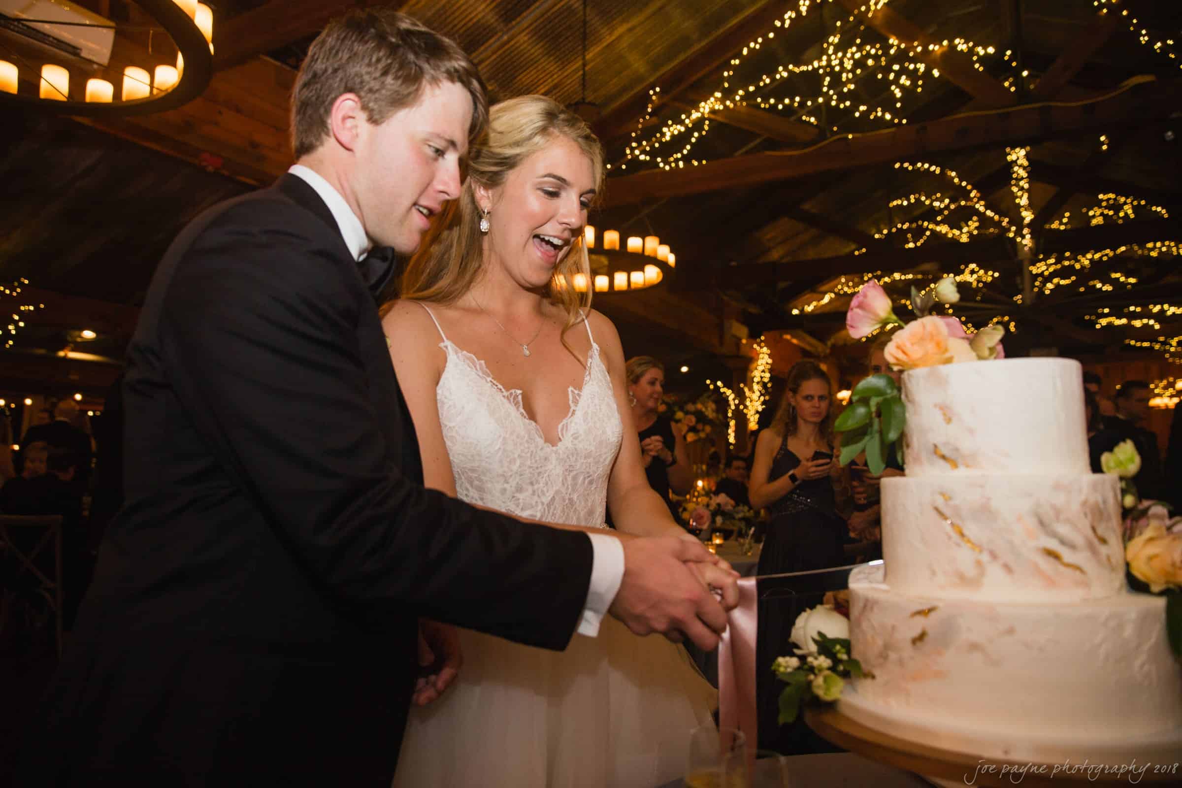 A bride and groom happily cut their wedding cake. The venue is warmly lit with string lights and chandeliers, creating a festive atmosphere. The cake is decorated with flowers. Guests are visible in the background.
