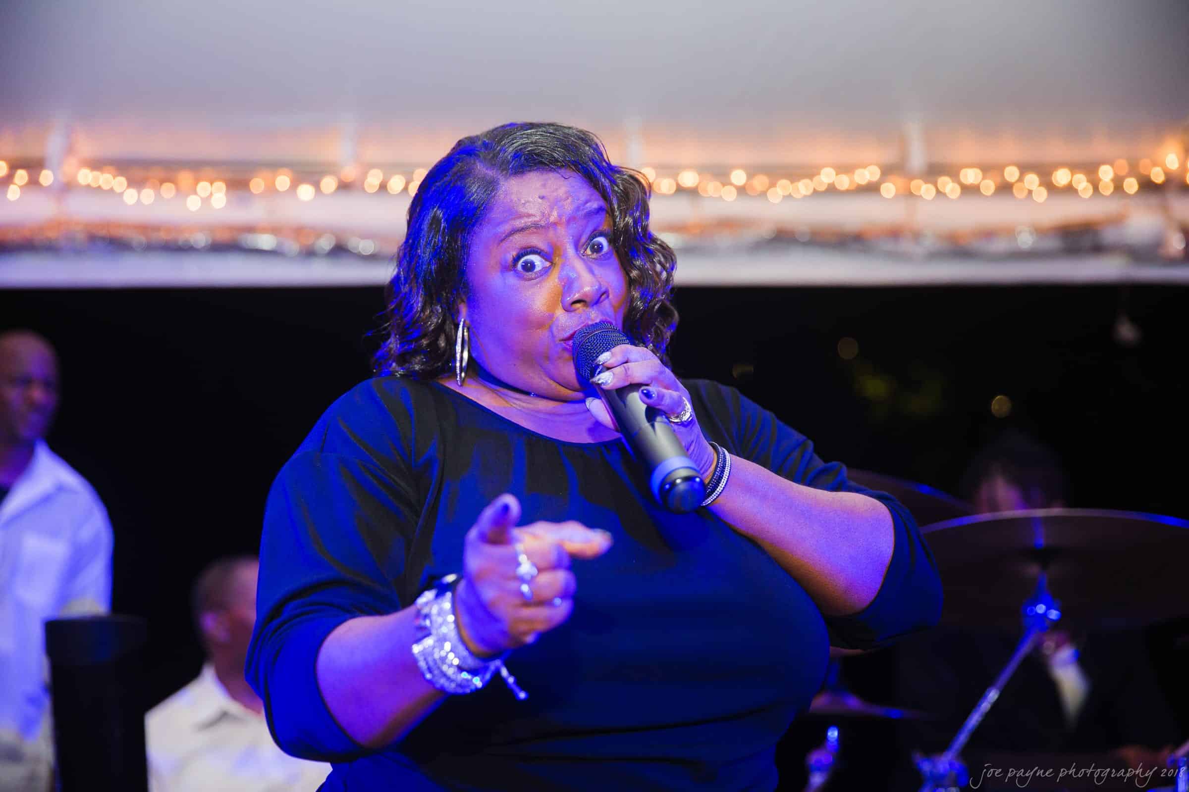 A woman passionately singing into a microphone, wearing a black outfit and accessorized with earrings and bracelets. She gestures forward, illuminated by stage lights with a backdrop of string lights.