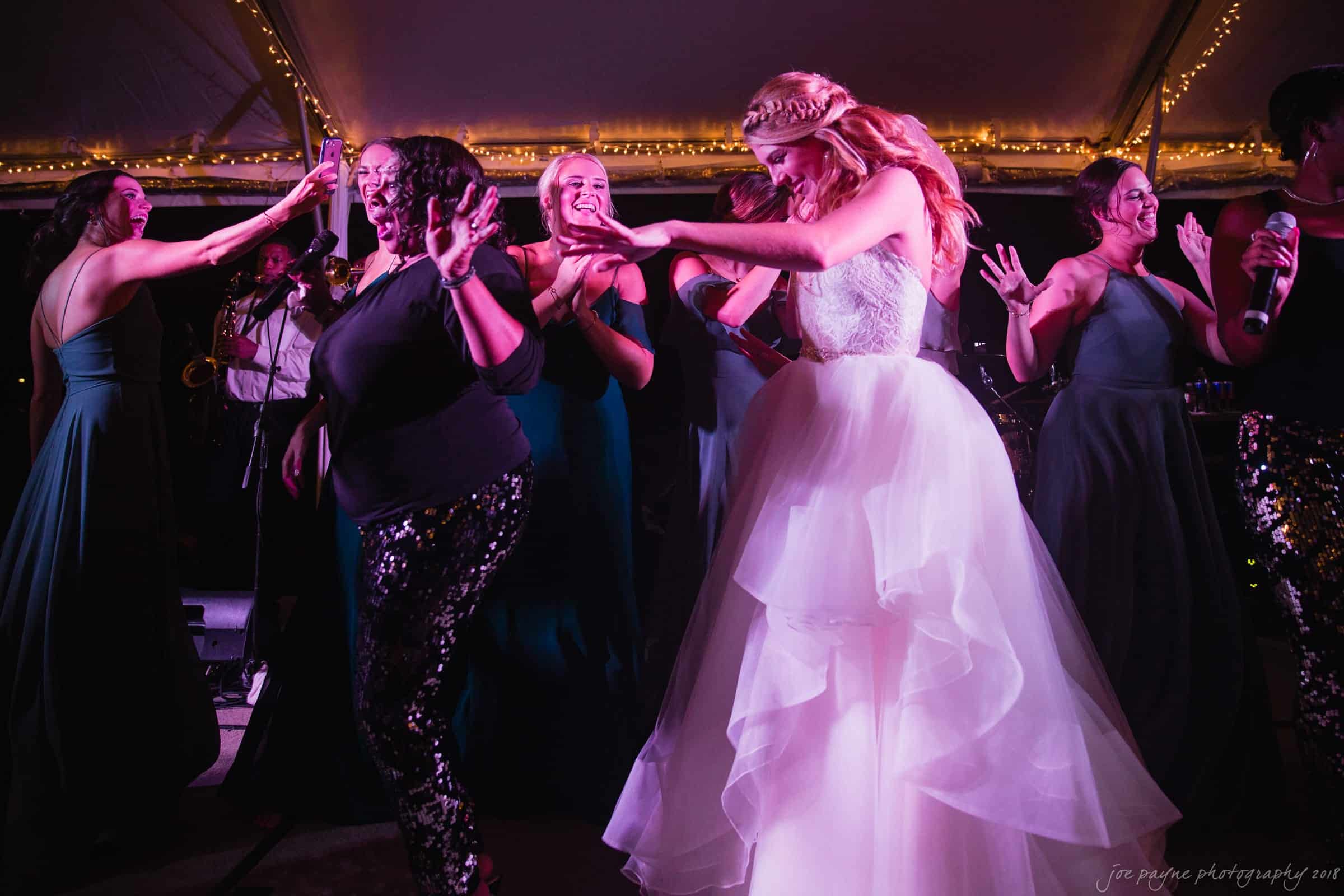 A bride in a white gown dances with bridesmaids and guests under a tent adorned with string lights. The energetic group is enjoying music, with some clapping and others laughing, creating a joyful atmosphere.
