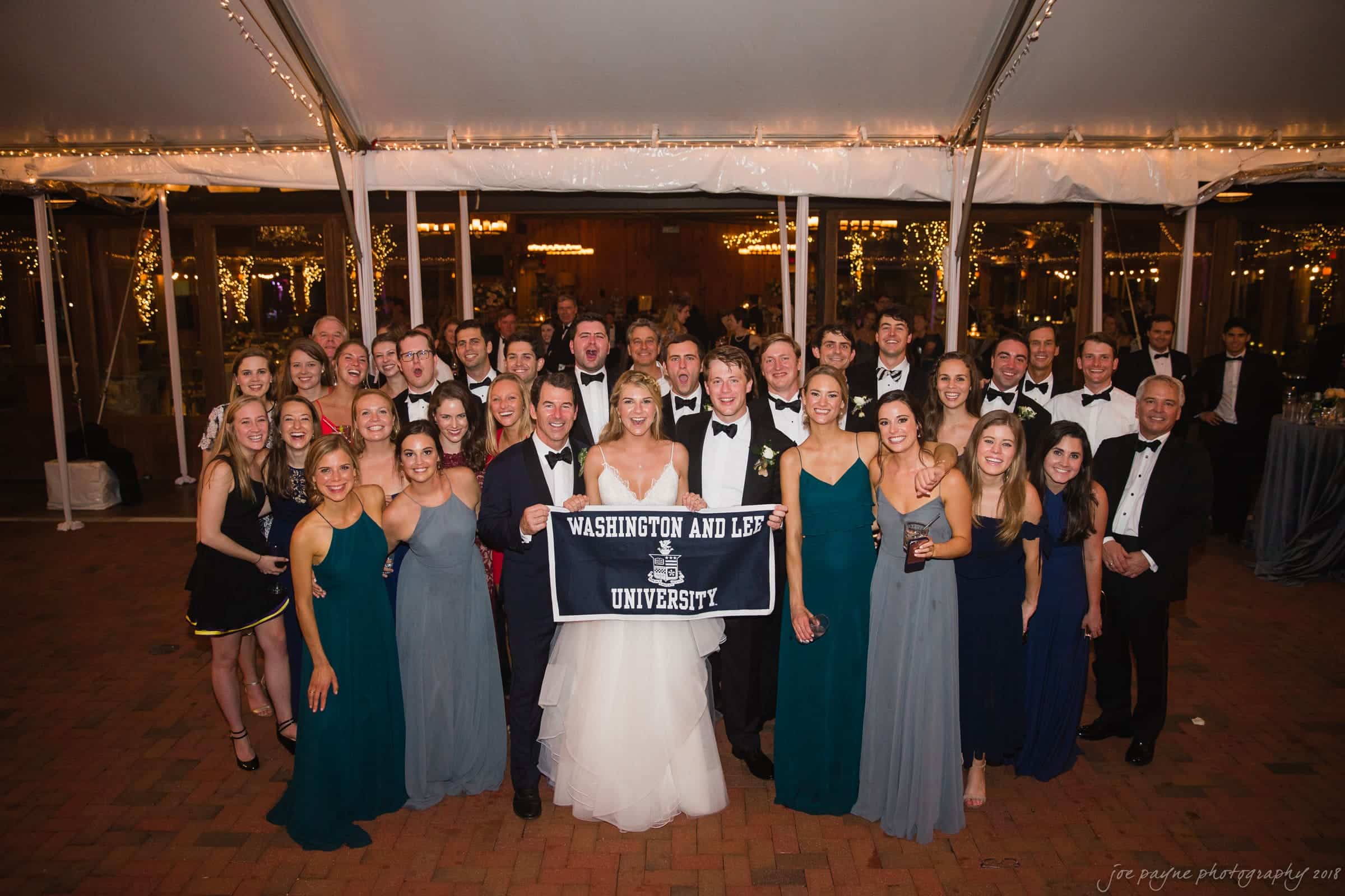 A large group of people dressed in formal attire poses under a tent. A couple in the center holds a Washington and Lee University banner. Twinkling lights decorate the background.