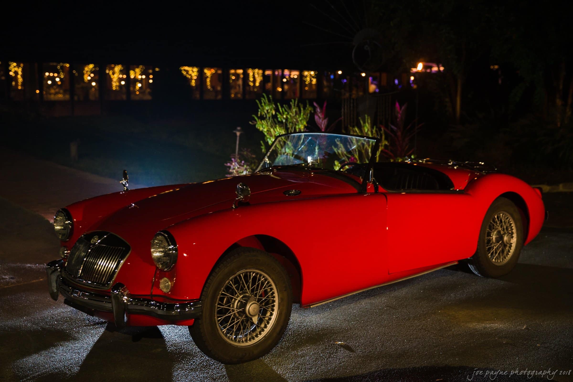 A classic red convertible sports car with wire-spoke wheels is parked outdoors at night. The vehicle is illuminated, with decorative lights visible in the background.