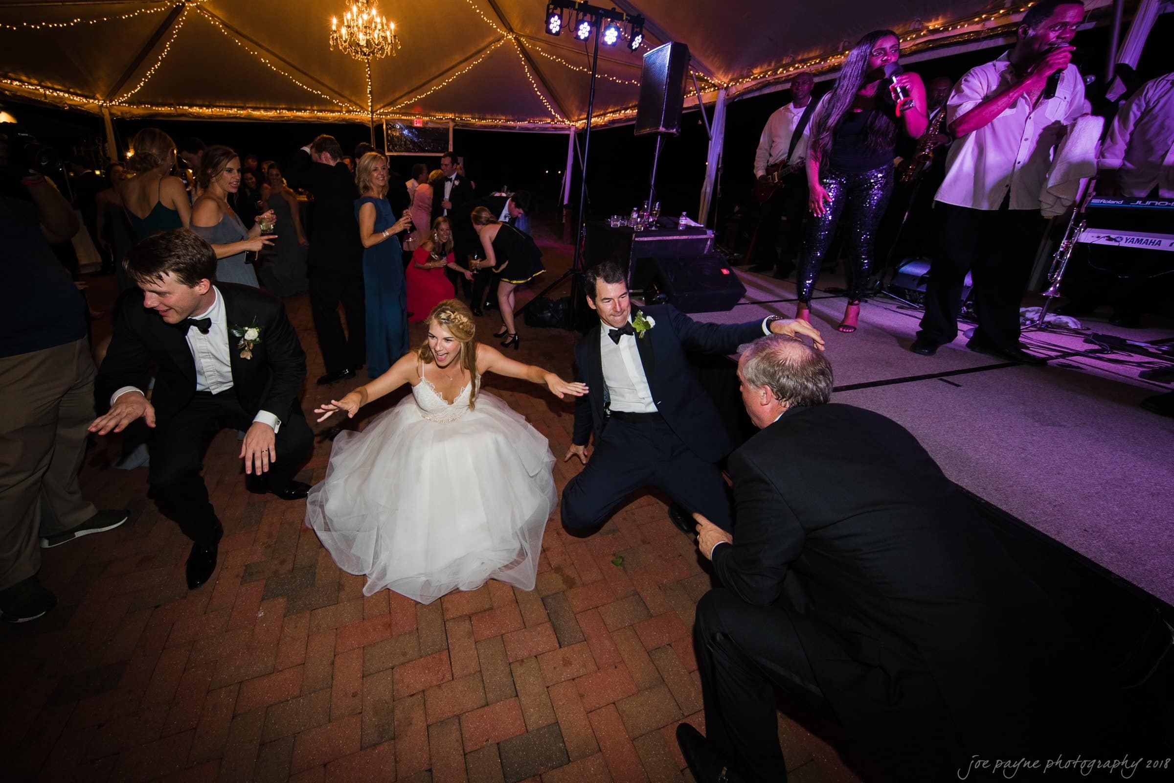 A bride and groom dance energetically on the floor with two men in suits at an outdoor nighttime wedding reception. Guests in the background enjoy the music played by a live band on stage under a tent with string lights.