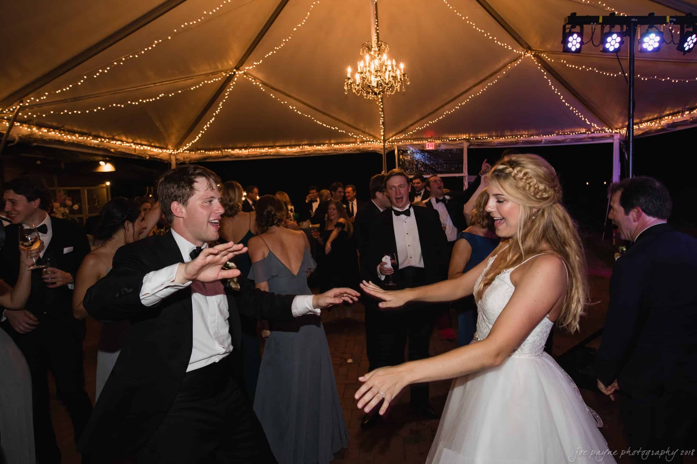 A couple dances energetically under an elegant, lit-up canopy at an outdoor evening event. The woman wears a white dress and the man is in a black tuxedo. Other guests in formal attire are visible in the background, enjoying the festive atmosphere.