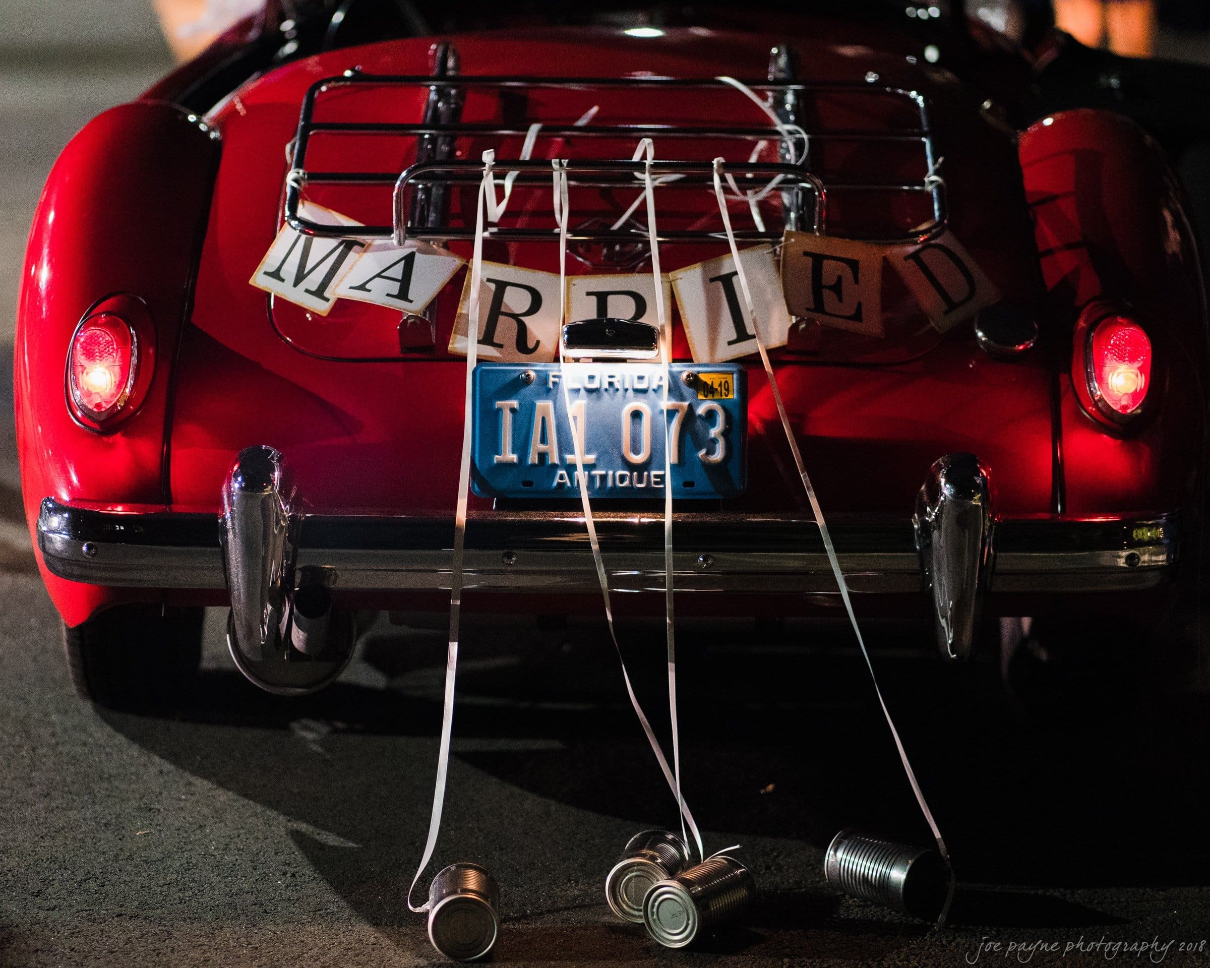 A vintage red convertible with a Just Married sign and cans tied to its bumper. Florida license plate reads 1AL 073. Its nighttime and the car is seen from the back.