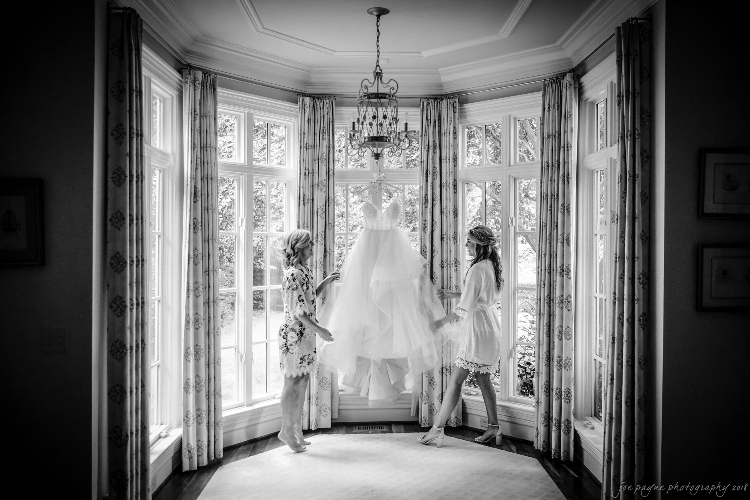 Two people stand by a large window with patterned curtains, holding a wedding dress. The scene is elegant and serene, set in a well-lit room with a chandelier above. The dress is prominently displayed in the center.