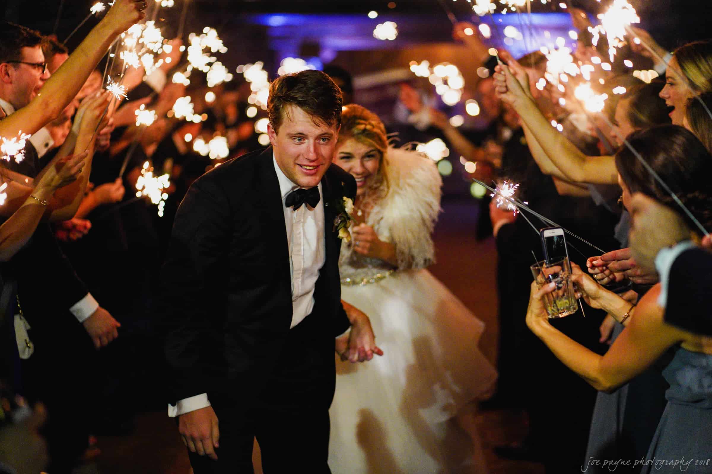 A bride and groom in formal attire walk through a joyful crowd holding sparklers at night. The bride wears a white dress with feather details, while the groom is in a tuxedo. The setting is festive and celebratory.