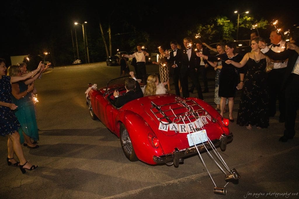 A newlywed couple in a red convertible drives away at night. The car has a Just Married sign and trailing tin cans. Friends and family stand on either side, holding sparklers and cheering in celebration.