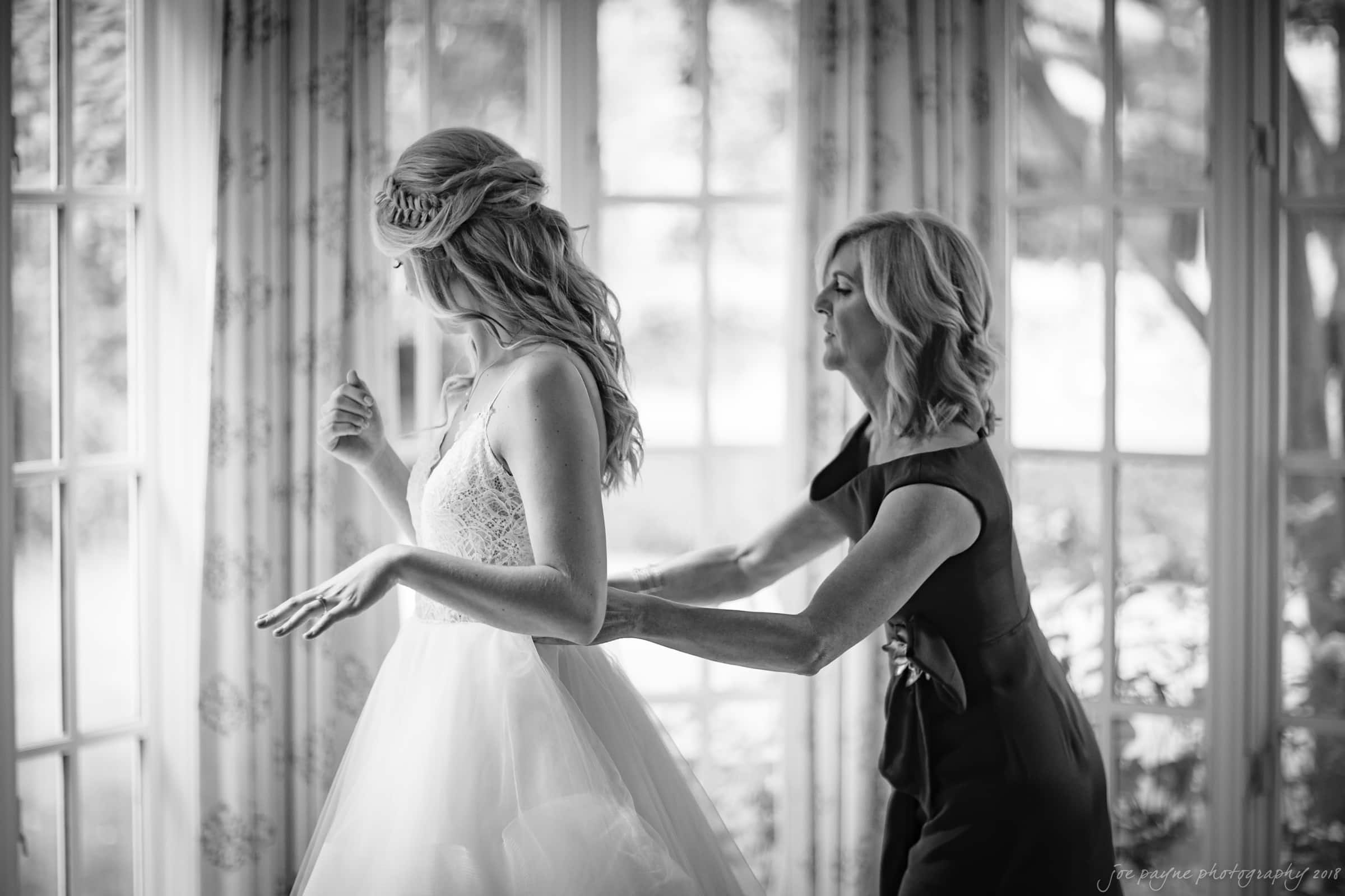 A black and white image shows a bride in a white gown with lace details standing by a window. She is being assisted by a woman in a dark dress who appears to be adjusting the gown. The ambiance is light and airy.