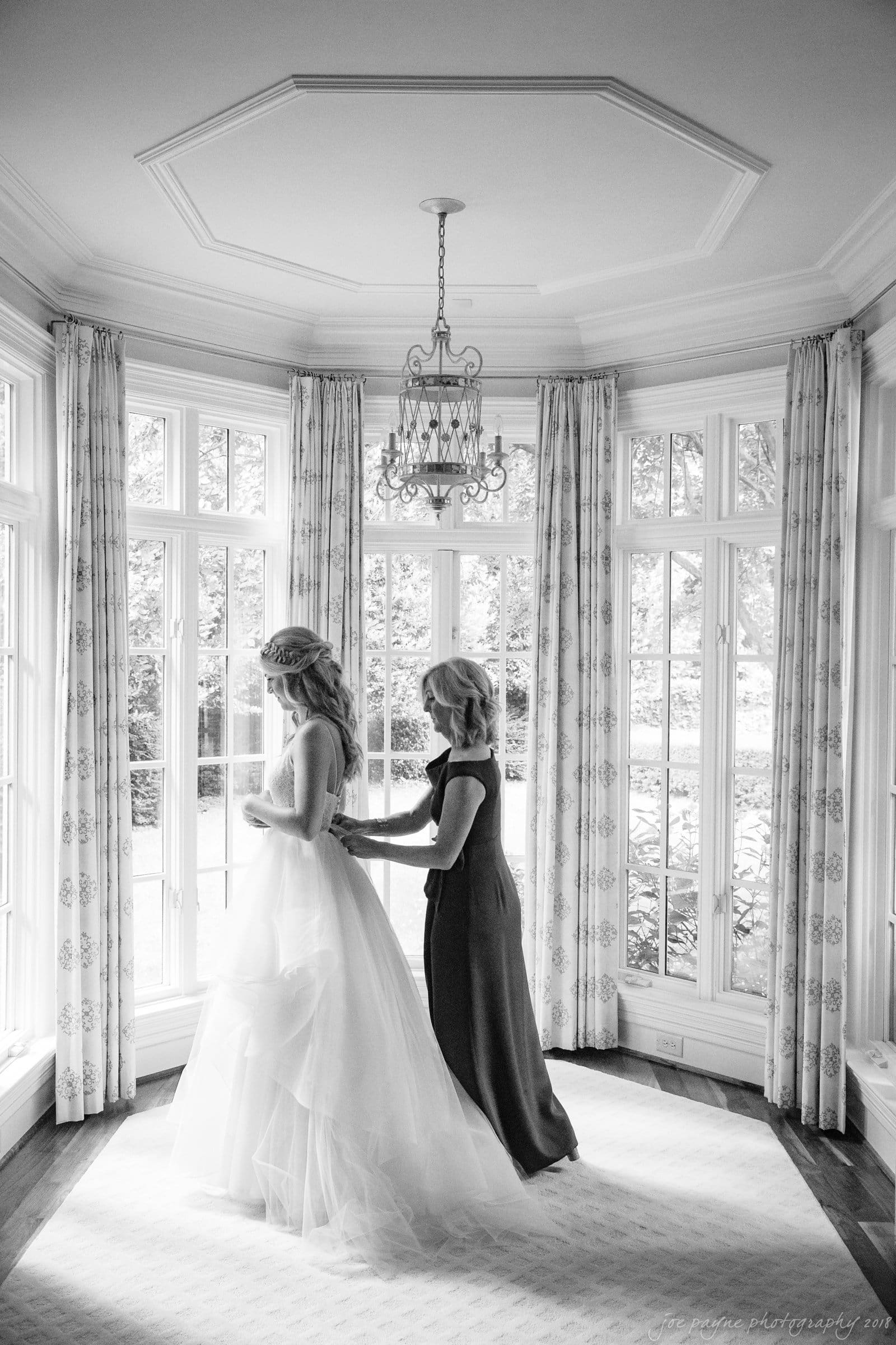 A bride in a white wedding dress stands in a bright room with large windows. Another woman, in a dark dress, adjusts the brides gown. They are under an ornate chandelier, with curtains draping the tall windows behind them.