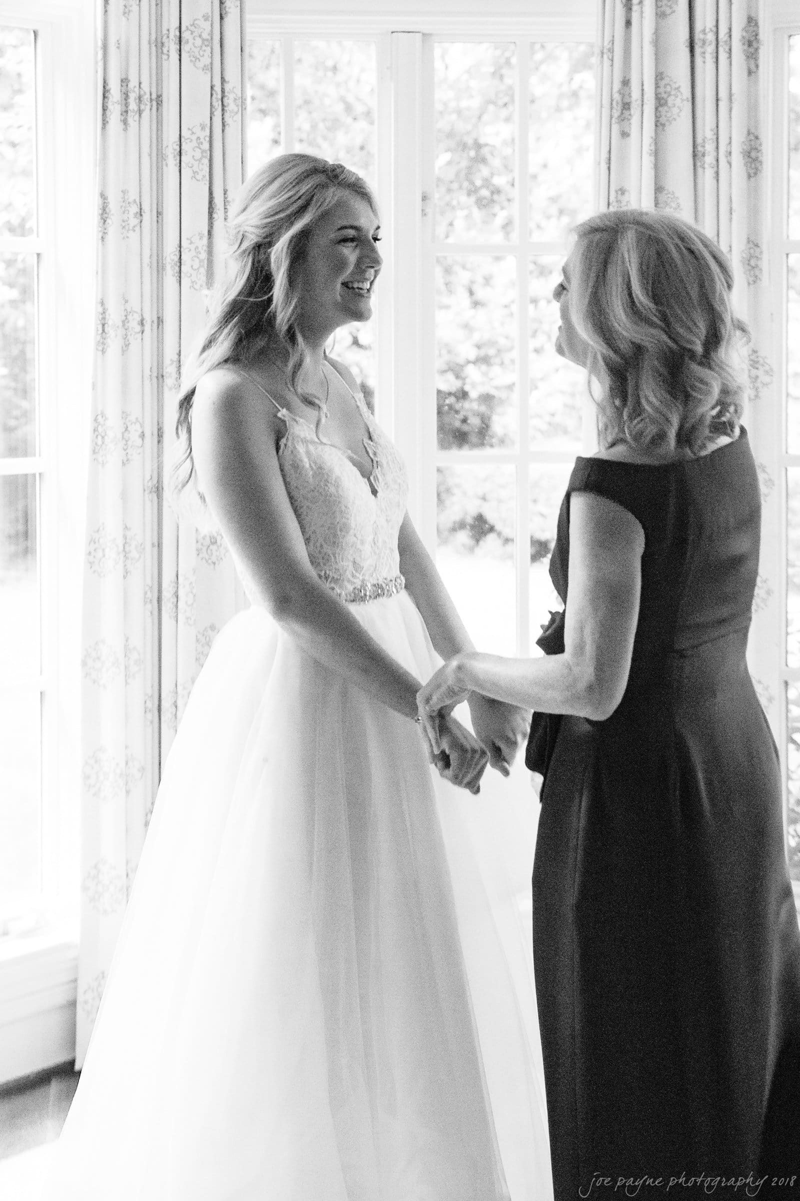 A bride in a white wedding dress smiles while holding hands with a woman in a dark dress. They stand near a large window, creating a softly lit, intimate moment.