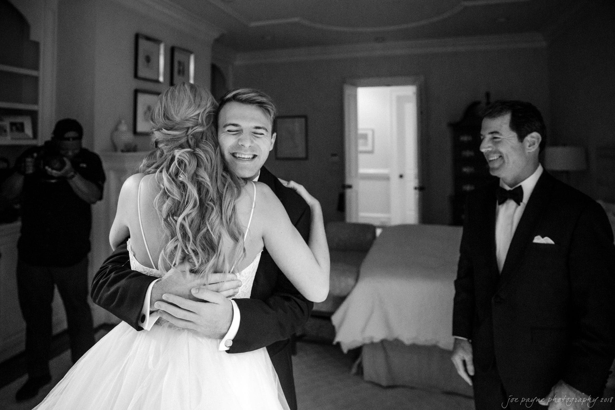 A bride in a white gown with loose curls hugs a young man in a suit, both smiling warmly. A man in a tuxedo stands nearby, smiling. The room is elegantly furnished with framed art on the wall and soft lighting.