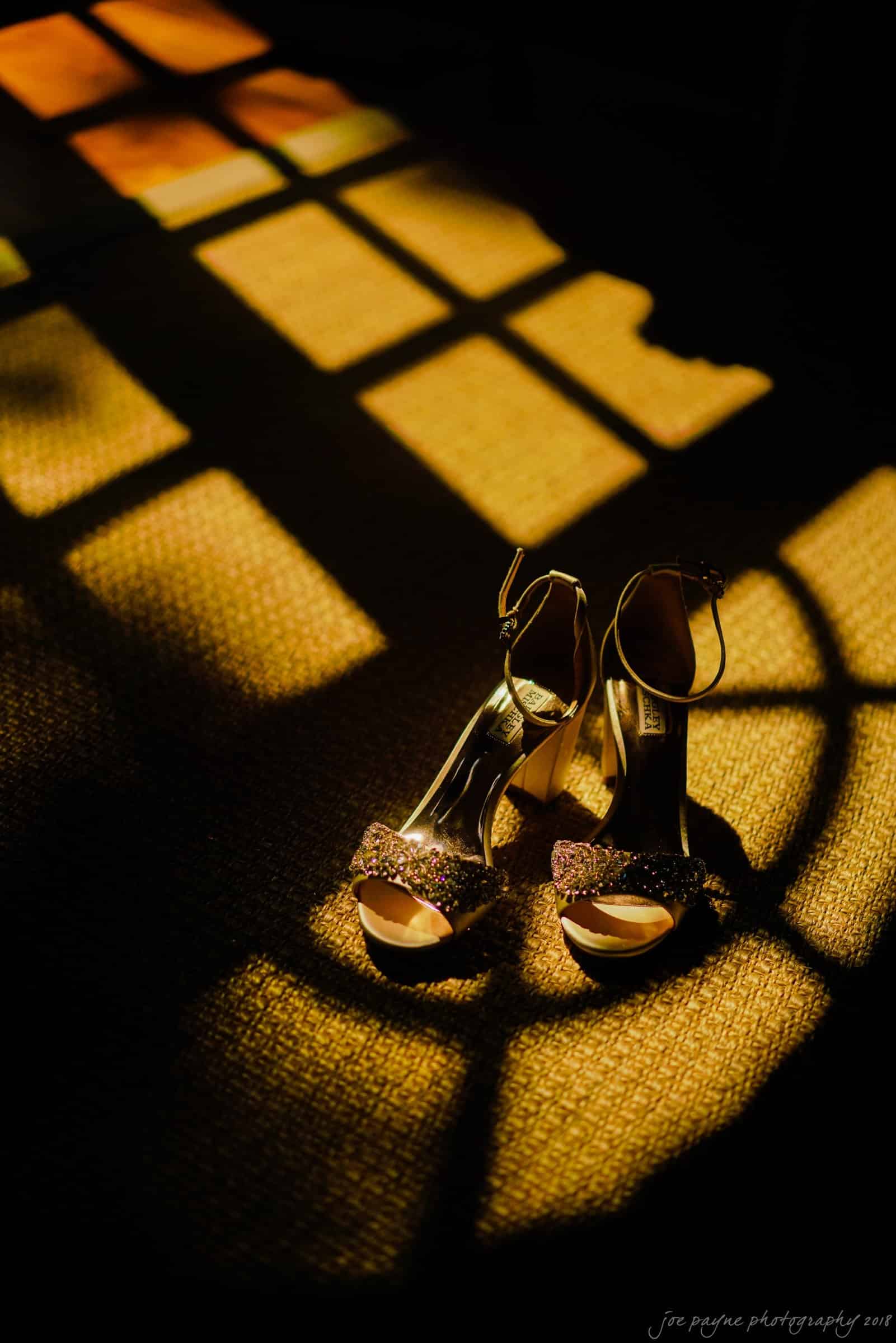 A pair of sparkling high-heeled shoes on a sunlit, textured carpet with shadow patterns from a nearby window.