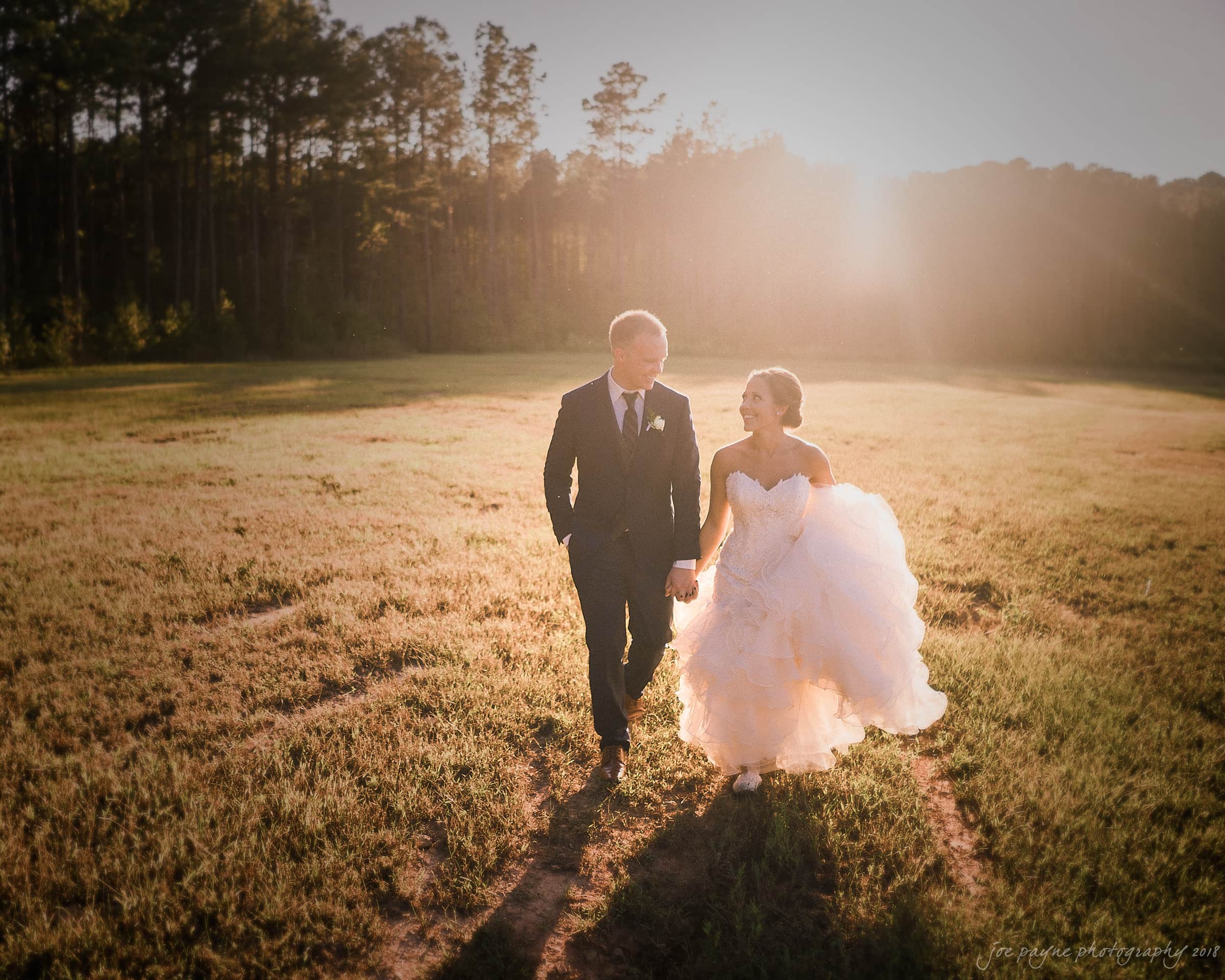 pavilion at carriage farm wedding - ashley & devon A bride and groom walk hand in hand across a sunlit field at sunset. The bride wears a flowing white gown, and the groom is in a dark suit. Tall trees border the field in the background, casting long shadows.