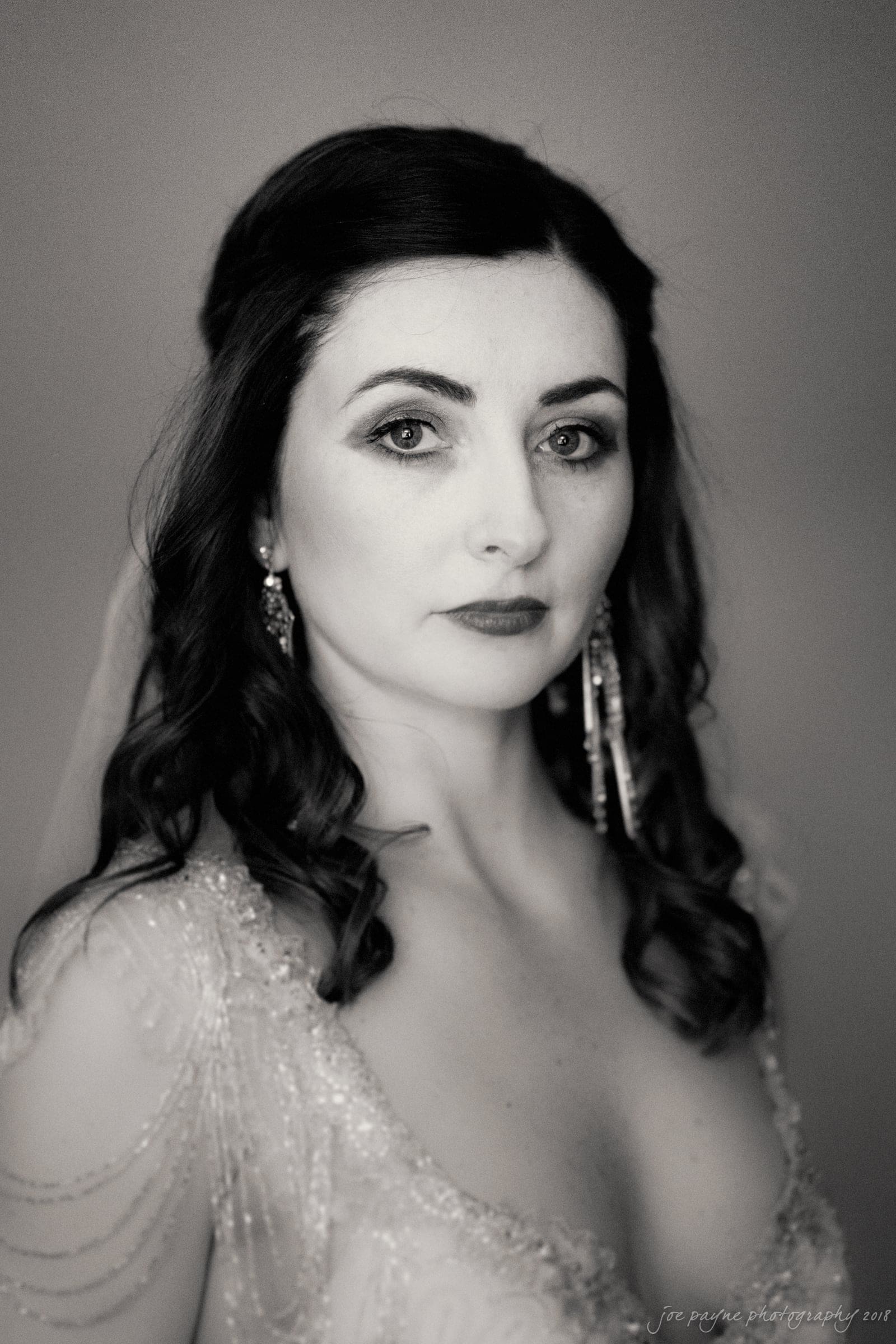 Black and white portrait of a woman with long, wavy hair wearing a detailed, beaded dress and long earrings. She has a neutral expression and is looking directly at the camera, with soft lighting highlighting her features.