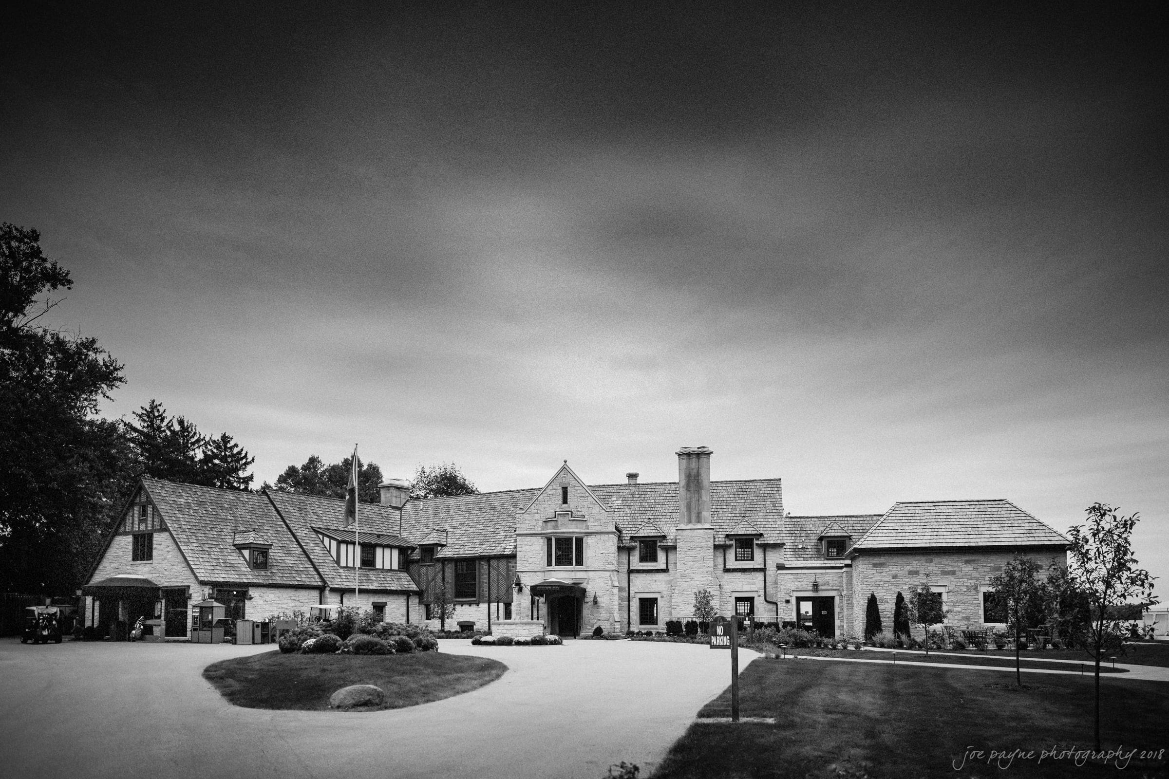 A large, elegant stone mansion with multiple chimneys and a circular driveway. The sky overhead is overcast, giving the black and white image a dramatic look. Lush trees and a manicured lawn surround the house.