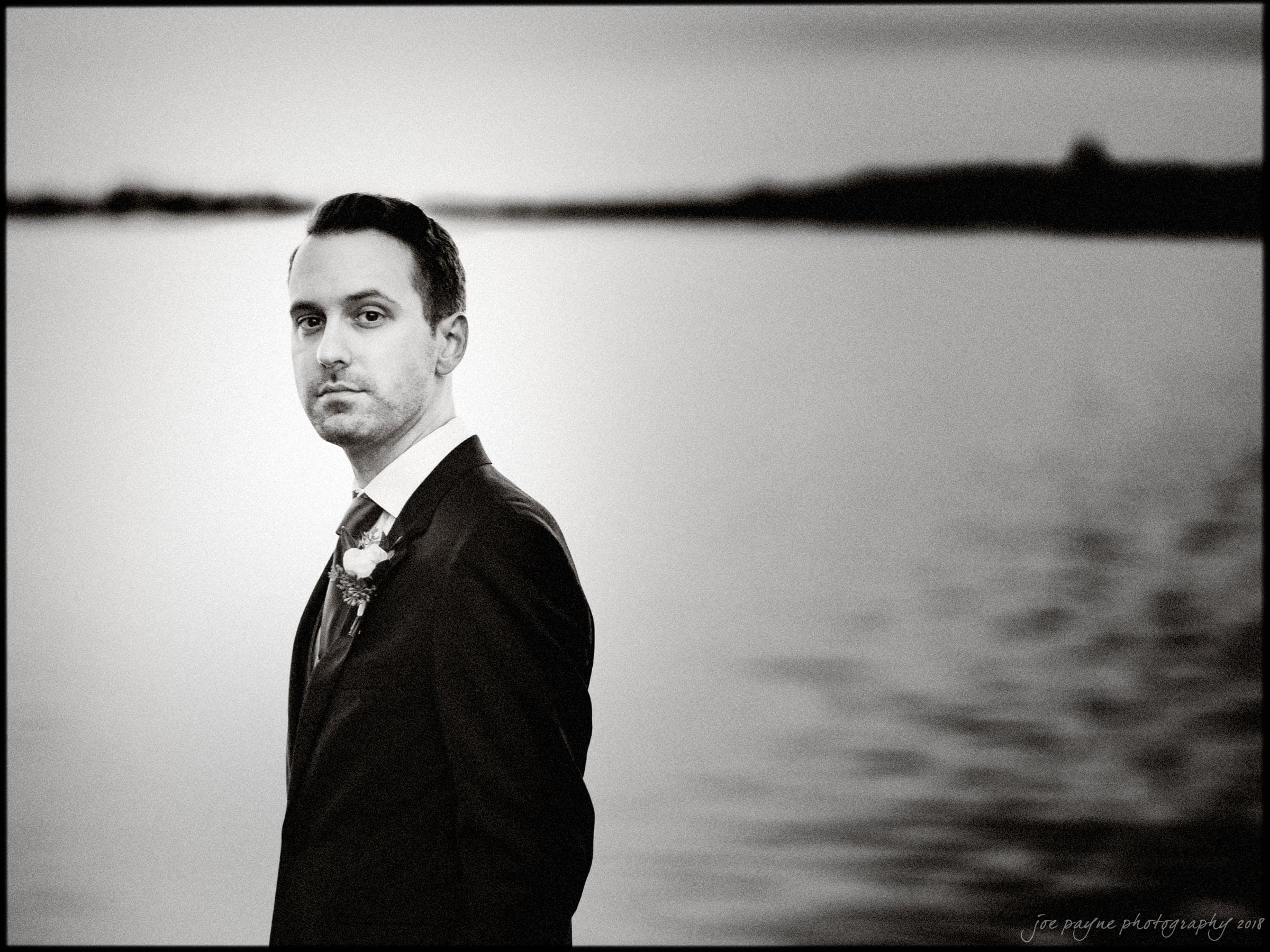 A man in a suit stands in front of a body of water, looking towards the camera. The photo is in black and white, with a blurred background that gives a serene and calm atmosphere.