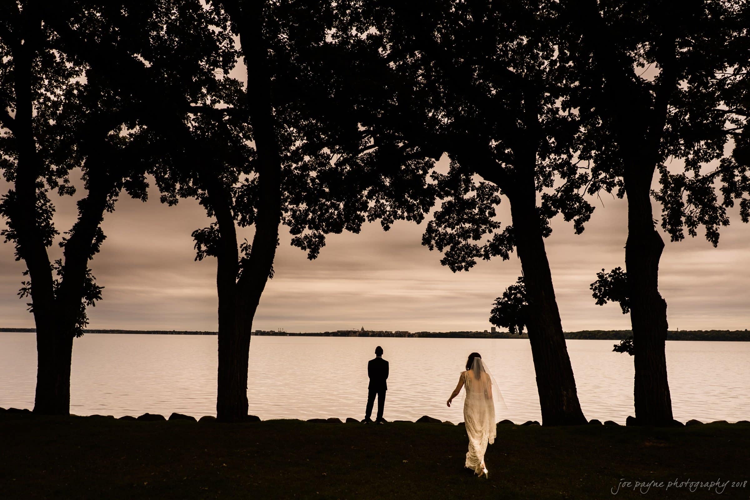 A silhouetted bride and groom stand apart between large trees, overlooking a calm lake under a cloudy sky. The bride, dressed in white, is walking towards the groom, who stands closer to the waters edge.