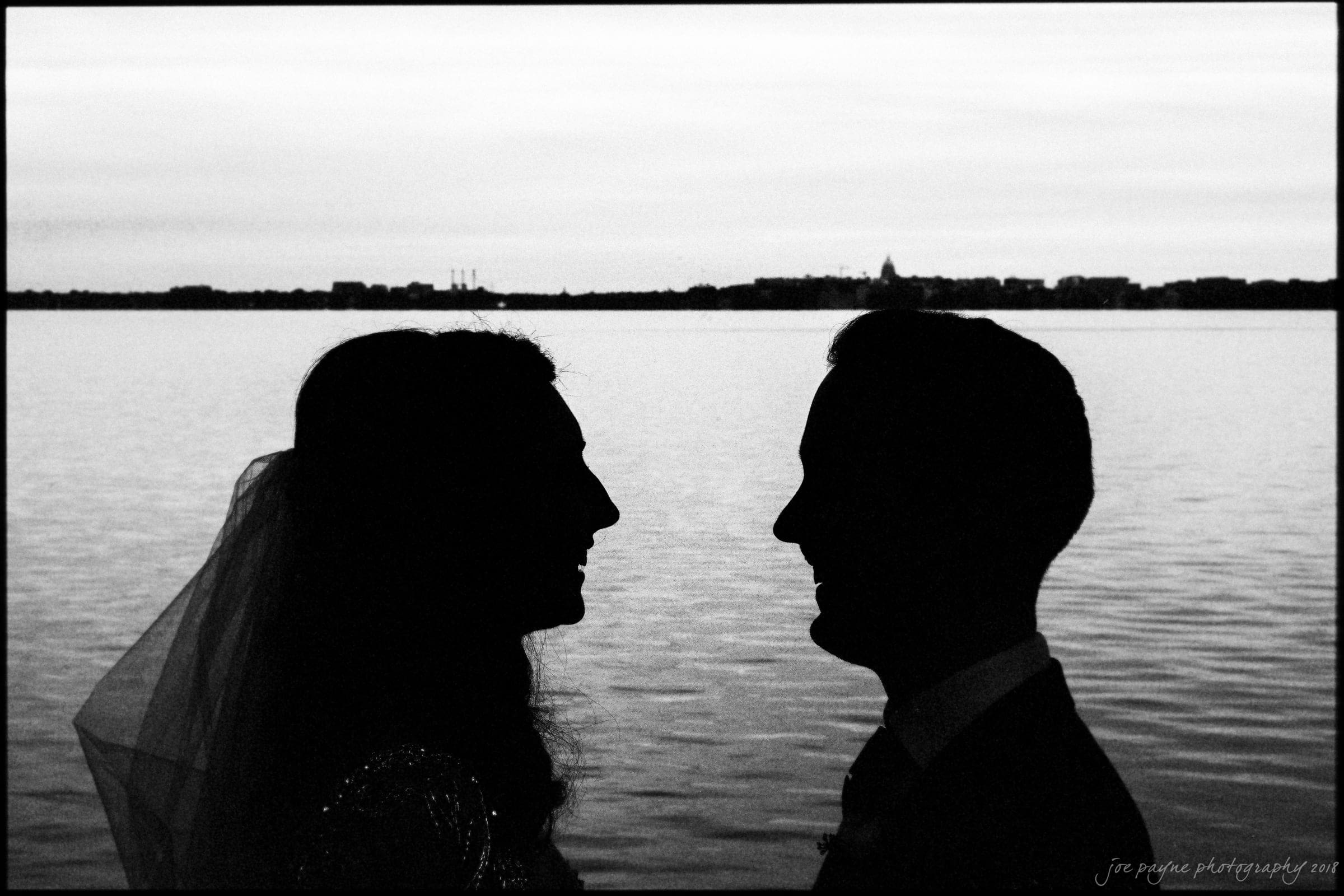 A silhouetted couple face each other against a tranquil waterfront backdrop. The evening sky and distant city skyline create a romantic atmosphere.