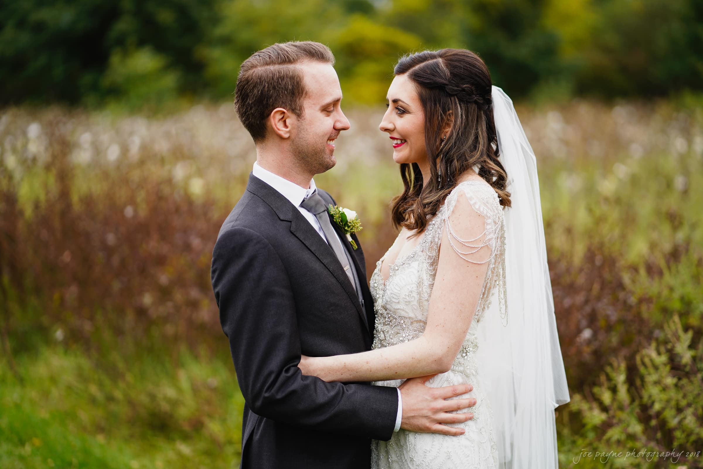 A bride and groom stand close together outdoors, smiling at each other. The bride is wearing a white lace dress and veil, while the groom is in a dark suit with a boutonniere. The background is a lush, green natural setting.