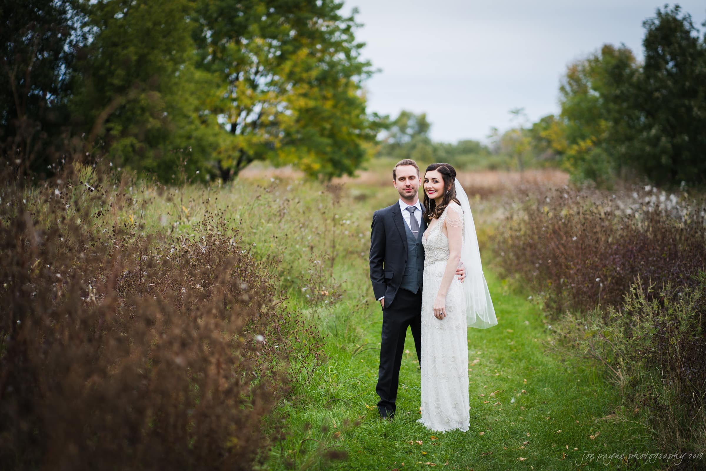 A bride in a white lace dress and veil stands beside a groom in a gray suit and purple tie. They are smiling in a grassy, overgrown field with trees and cloudy skies in the background, capturing a serene and romantic moment.