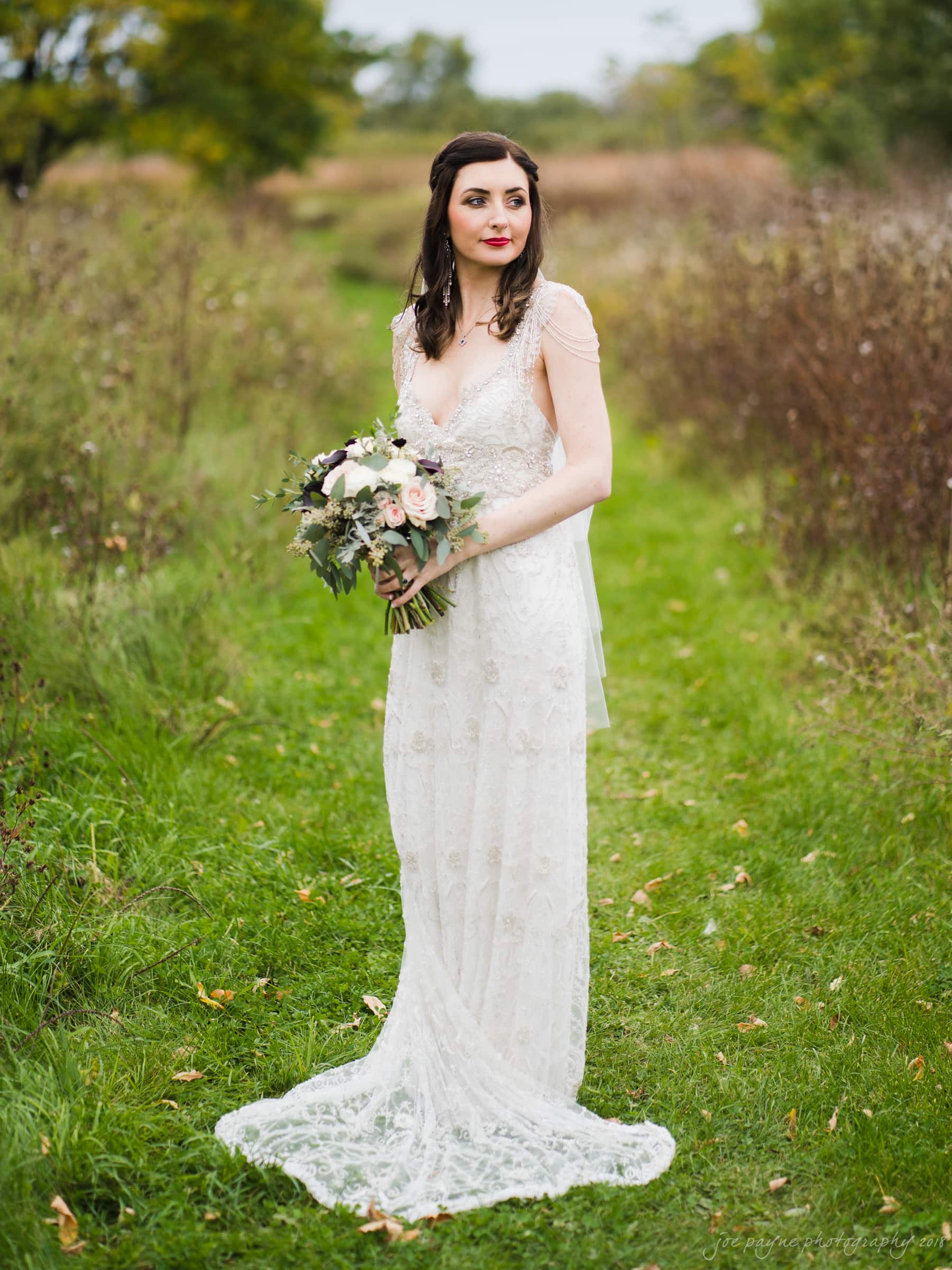 A bride in a white lace gown holds a bouquet of flowers, standing on a grassy path surrounded by trees and shrubs. The scene is outdoors, with a natural, serene backdrop.