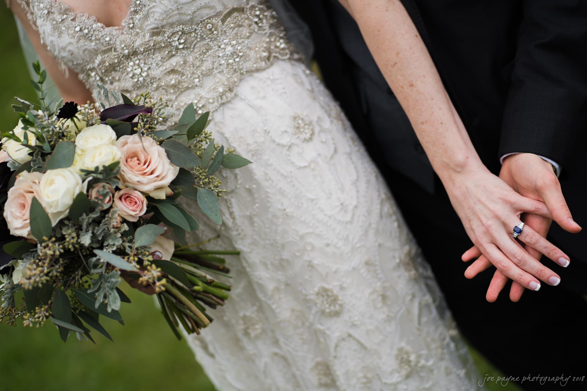 A bride and groom stand close, with entwined hands and rings visible. The bride holds a bouquet of roses and greenery, wearing a lace-embellished gown. The background is a grassy area, adding a natural touch to the scene.