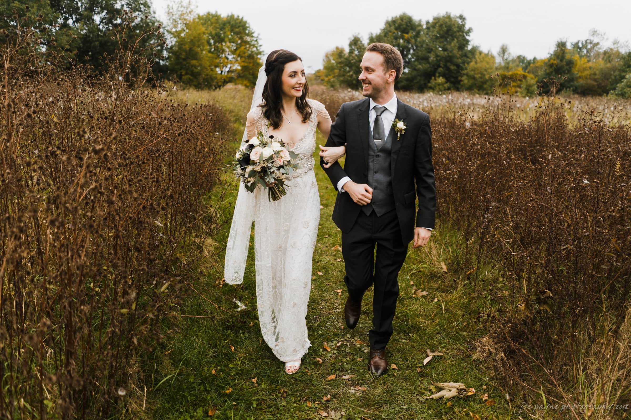 A bride in a white gown and a groom in a black suit walk arm in arm through a field. She holds a bouquet of flowers, and both are smiling. Tall grasses line the path, with trees in the background under a cloudy sky.
