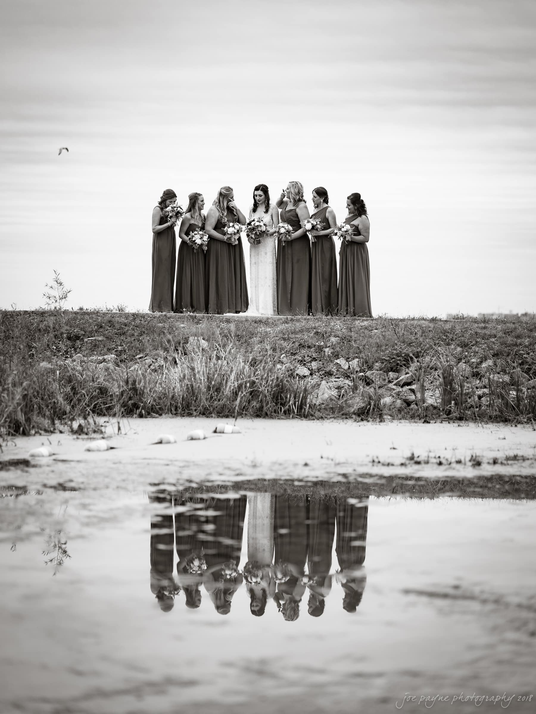 A bride and eight bridesmaids stand together on a grassy hill, holding bouquets. Their reflection is visible in a calm body of water below, with a cloudy sky in the background. The photo is in black and white.