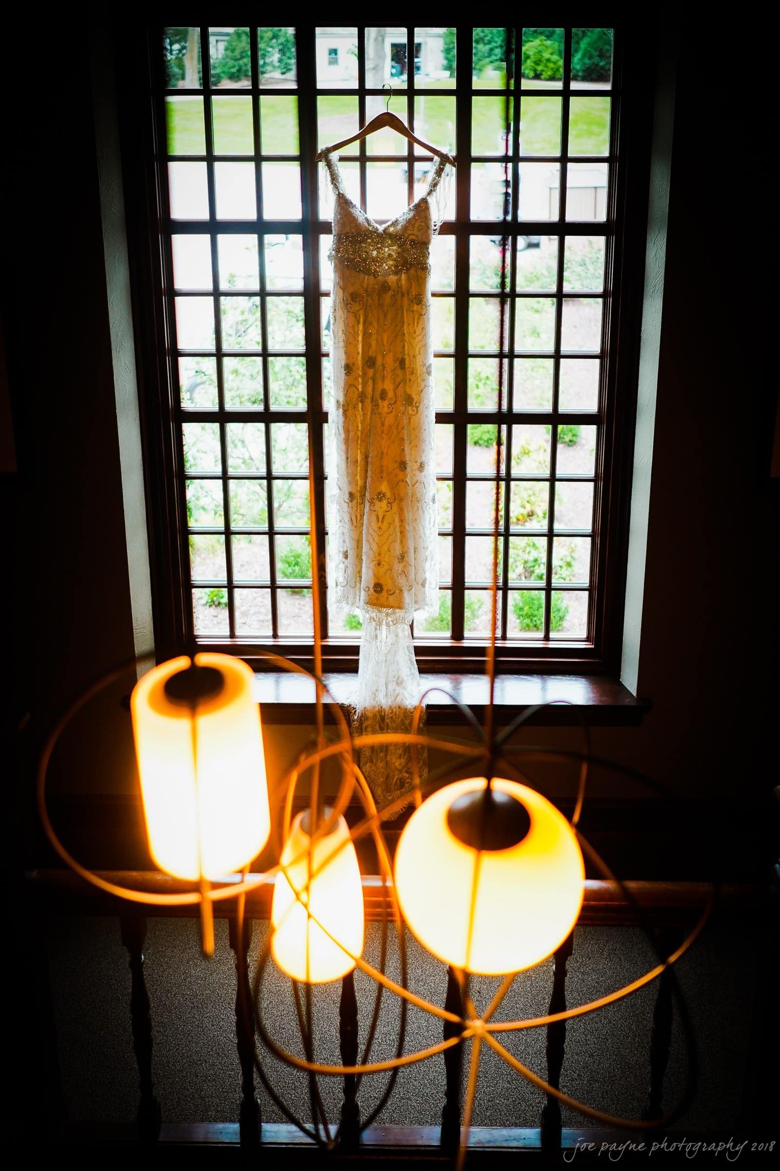 A delicate white wedding dress hangs in front of a large window with a grid pattern. The view outside shows greenery. Warm light from a modern chandelier illuminates the scene.