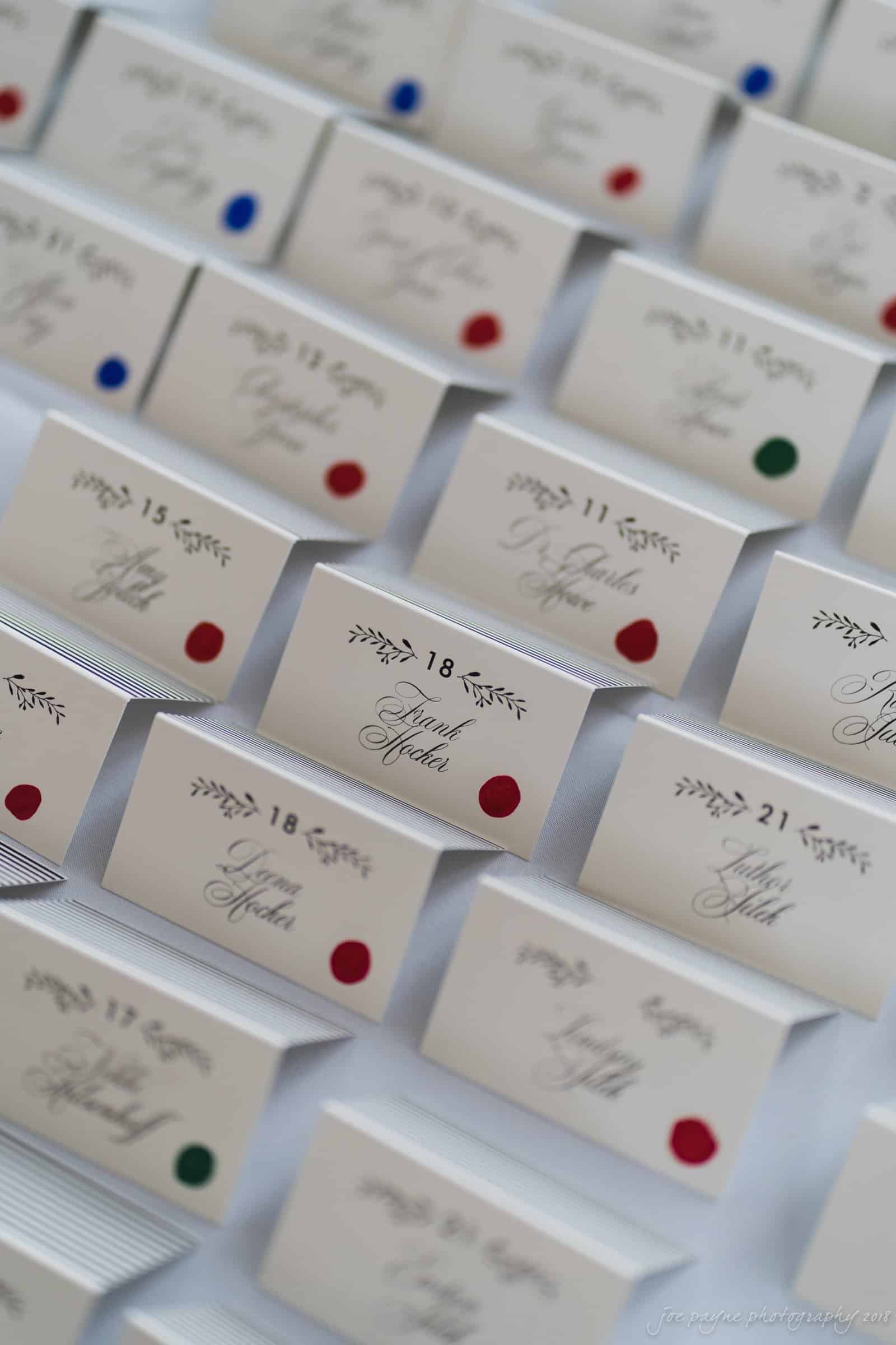 A display of elegant place cards arranged in rows. The cards have floral designs and names written in script, each marked with a different colored dot, including red, blue, and green. Some cards are numbered at the top.