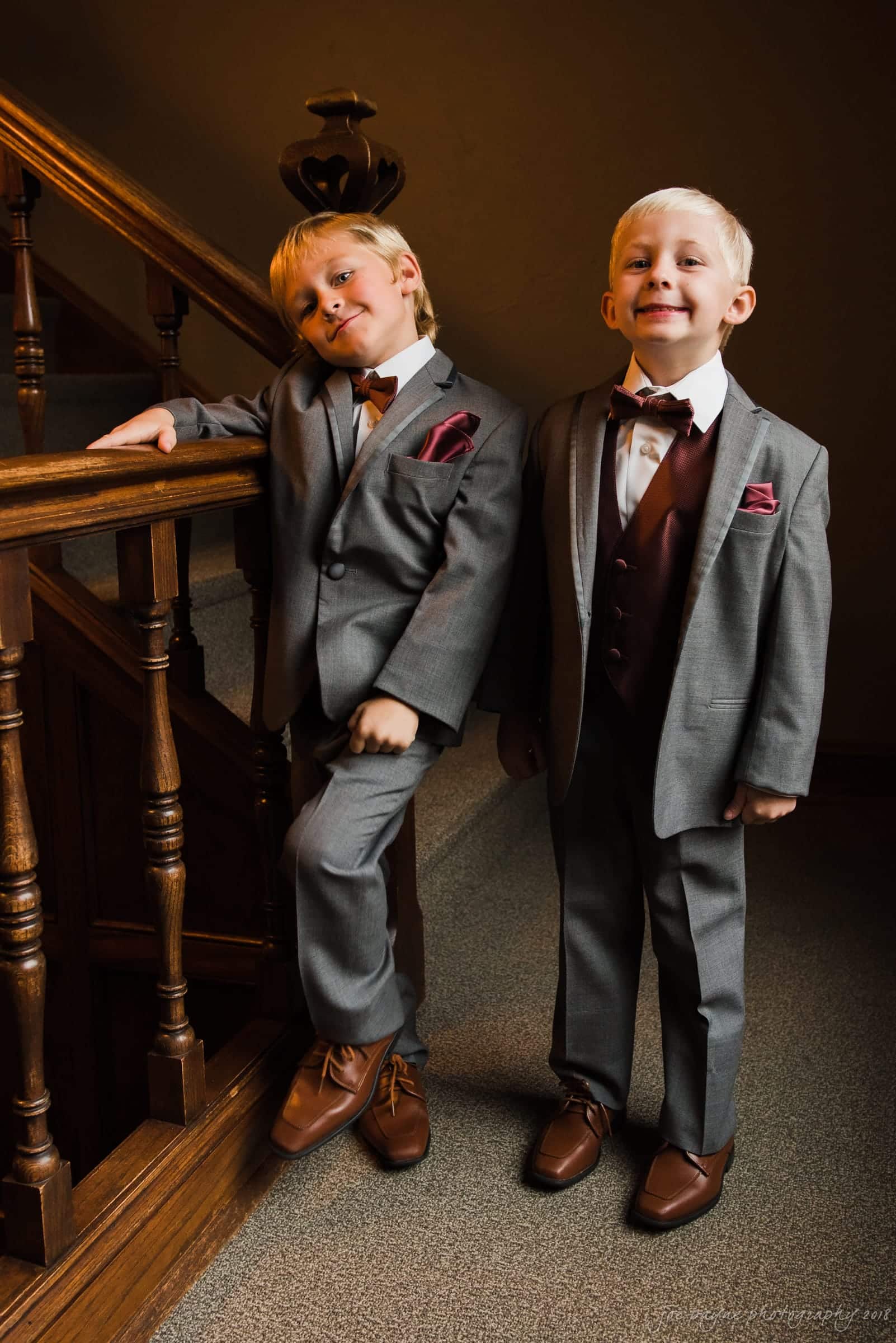 Two young boys in gray suits with burgundy vests and bow ties stand on a staircase. One leans casually against the railing, while the other stands upright, both smiling. The setting is indoors with wooden details.